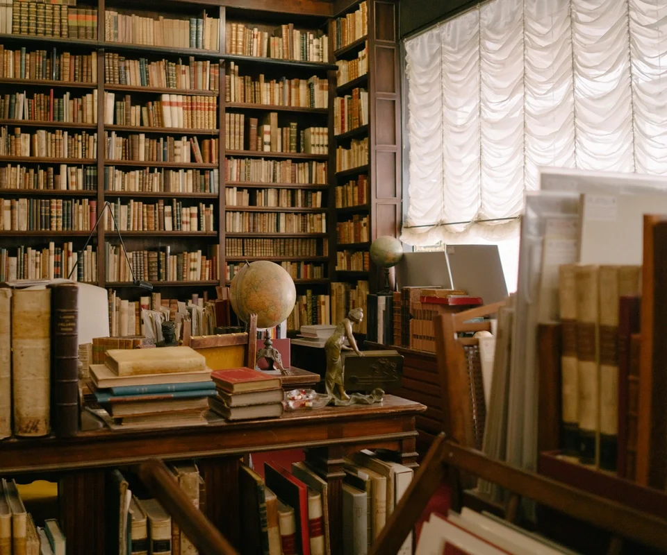 A room filled with tall wooden bookshelves packed with books, a large cluttered desk with many books, globes, and decorative items, and a window covered by draped curtains letting in diffused natural light.
