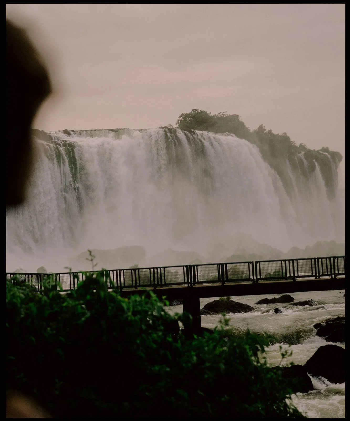 View of waterfalls with rushing water, mist, and a viewing platform in the foreground, surrounded by lush greenery.