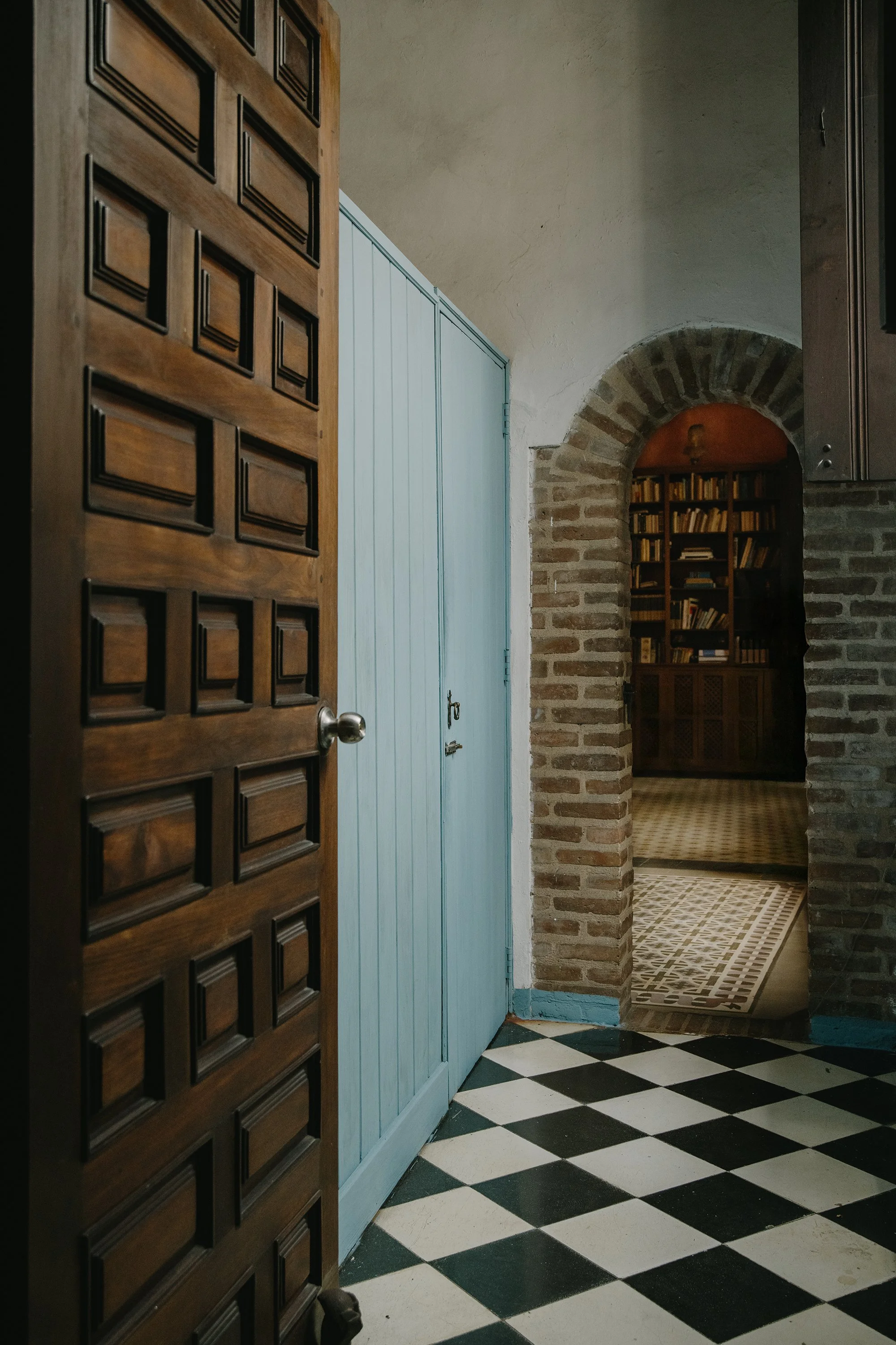 Interior view of a room with a wooden door partially open, black and white checkered floor, blue wall, brick archway leading to a bookshelf-filled room, and a small rug. Mundos Interiores. Ciudad Colonial de Santo Domingo.