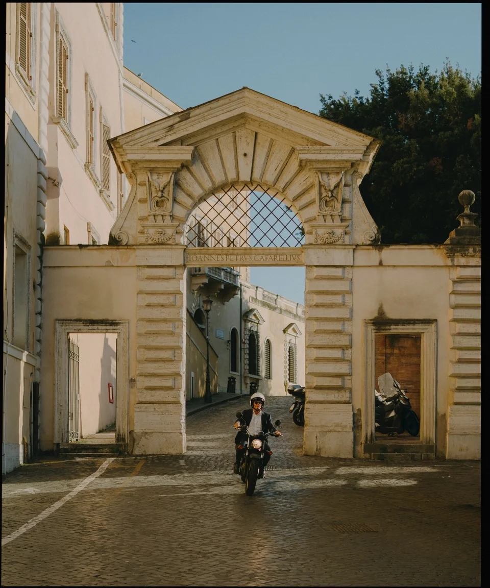 A person riding a motorcycle through an ornate archway in a European city, with cobblestone streets and historic buildings on both sides.