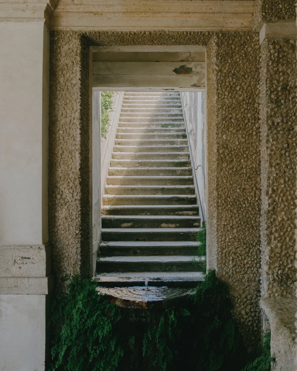 Outdoor stone staircase viewed through a rectangular opening in a textured wall, with water and greenery at the base.