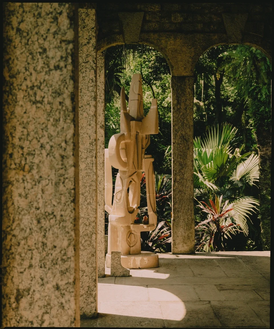 Stone sculpture of a geometric abstract figure on a patio with tropical plants in the background.