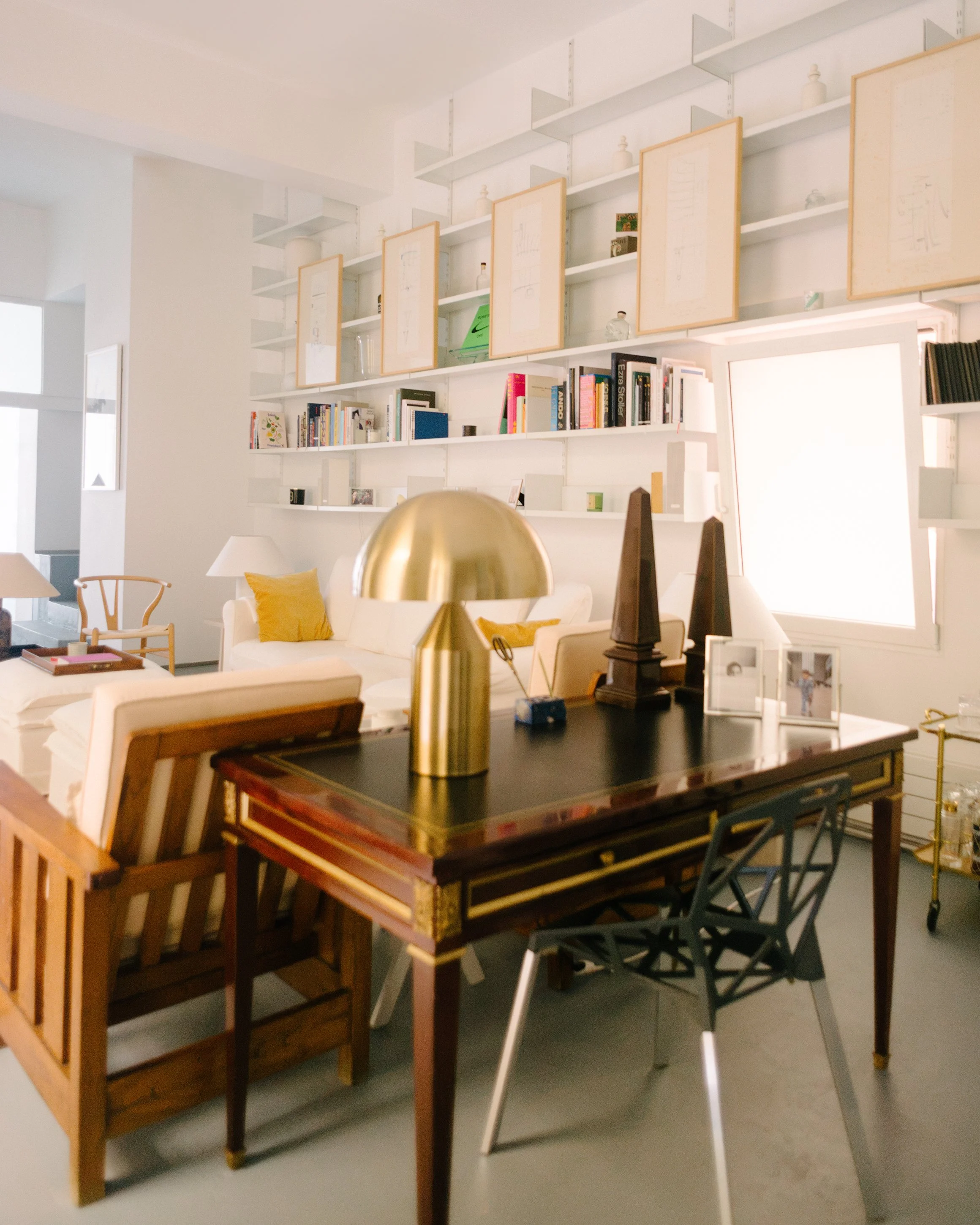 A living room with open white shelves mounted on the wall, displaying framed artwork, books, decorative objects, and a small plant. There is a white sofa with yellow pillows, a side table with a white lamp, and a table with framed photographs and a l