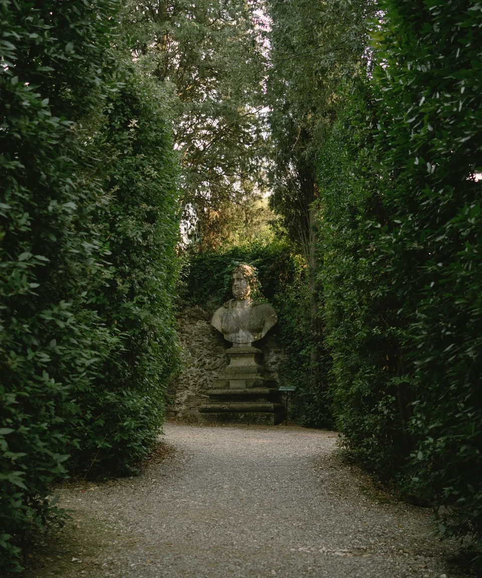 A stone bust of a man on a pedestal, situated outdoors amidst tall, dense green bushes and trees in a garden or park setting.