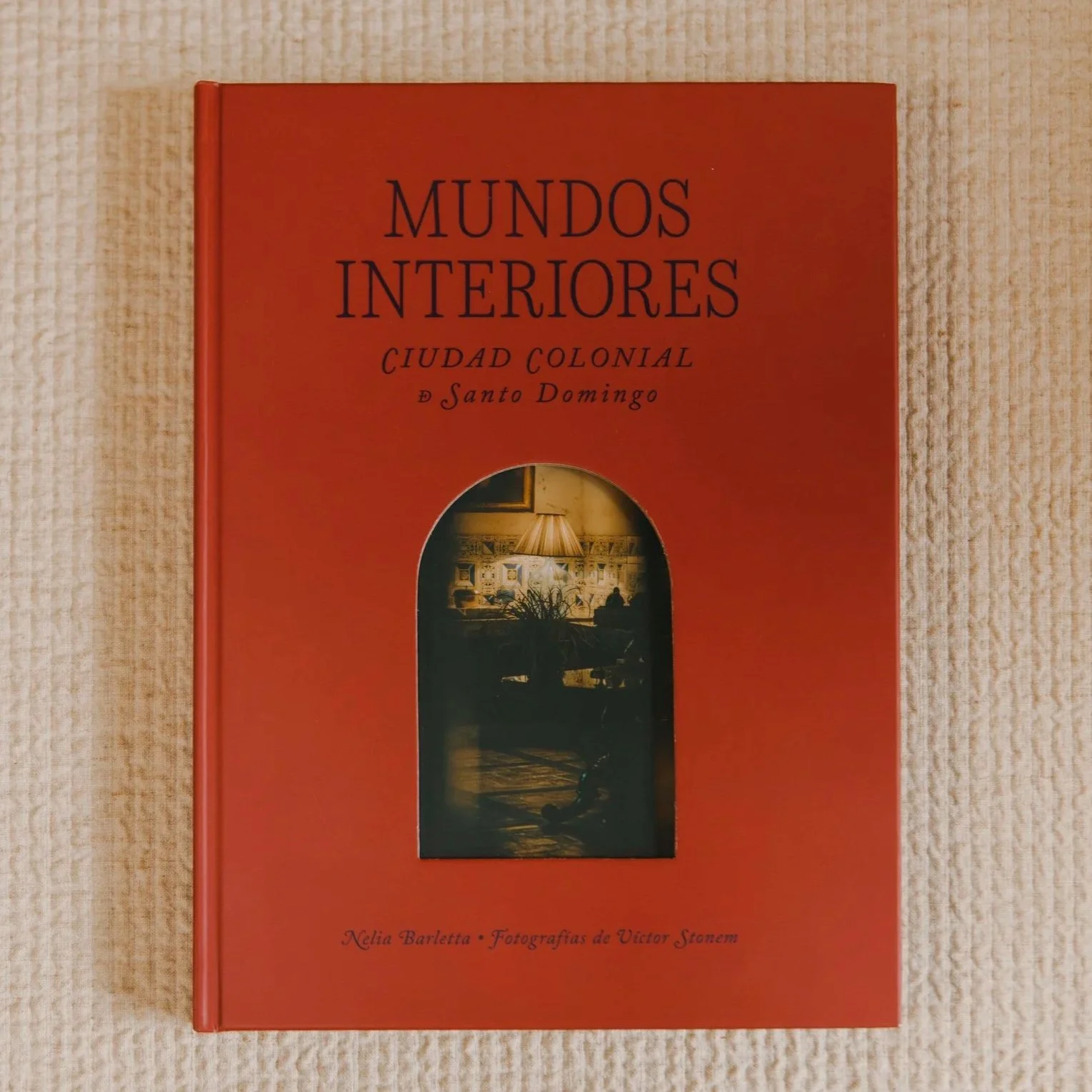 Red hardcover book titled "MUNDOS INTERIORES CIUDAD COLONIAL D Santo Domingo" with a small window revealing an interior scene with a lamp and furniture, resting on a textured beige surface.