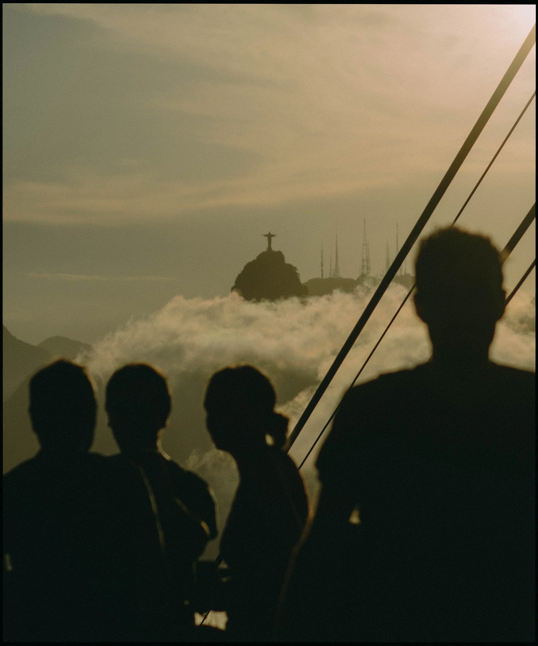 Silhouettes of people in the foreground with the Christ the Redeemer statue visible on a hill in Rio de Janeiro in the background, during sunset or sunrise.