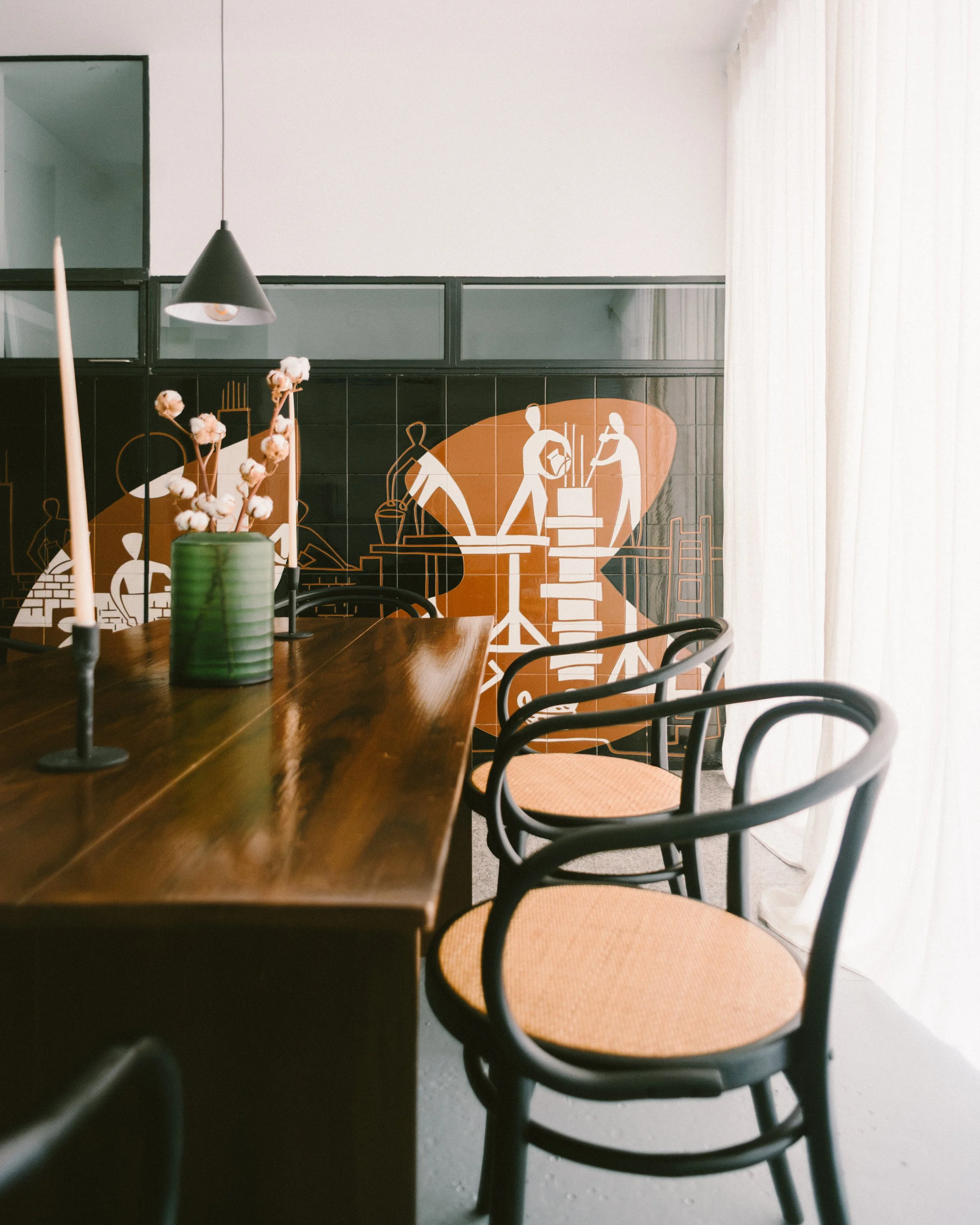 A dining room with wooden tables, black chairs with curved backs, a black pendant light, and a decorative wall art featuring illustrated workers and tools, with a window covered by light curtains on the right.