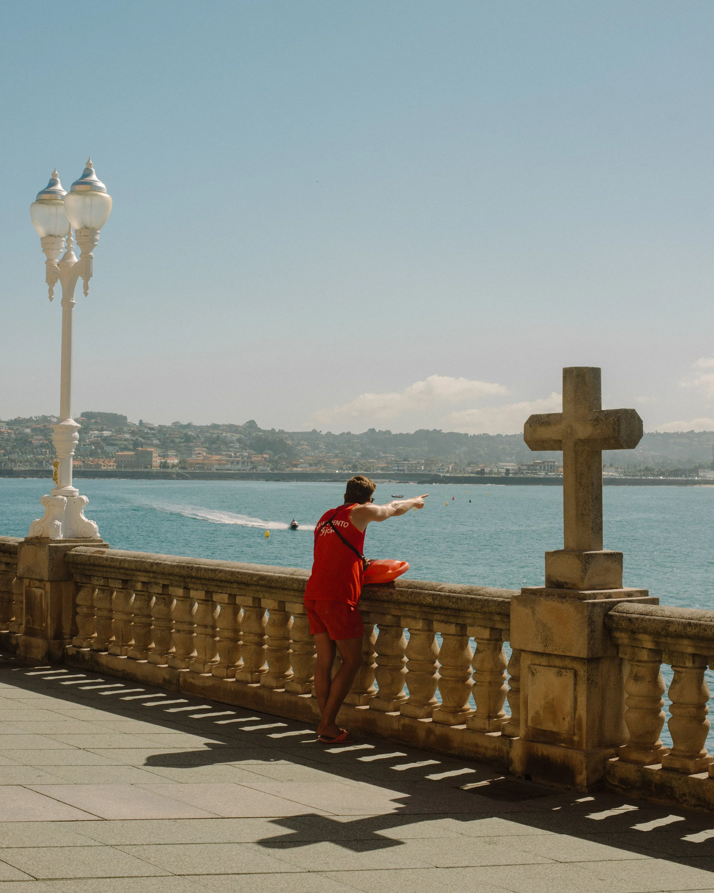 Person wearing red shorts and an orange tank top, leaning on a decorative stone railing by the water, pointing towards the sea, with a large cross monument in the background, under a clear sky