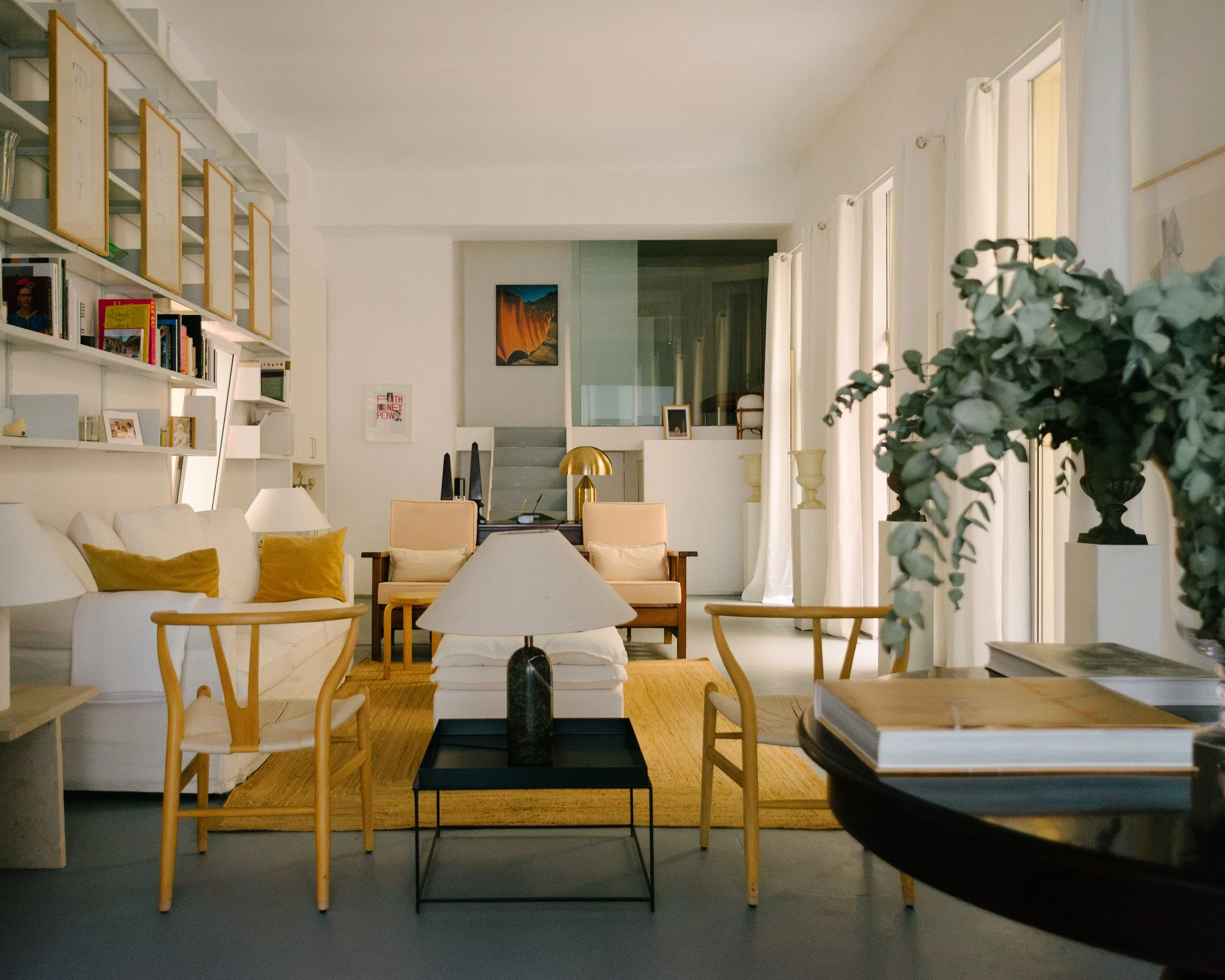 Bright living room with white walls and ceiling, featuring a white sofa with gold pillows, wooden chairs, a white bookshelf filled with books, and a large window with white curtains letting in natural light. There are decorative vases, framed picture