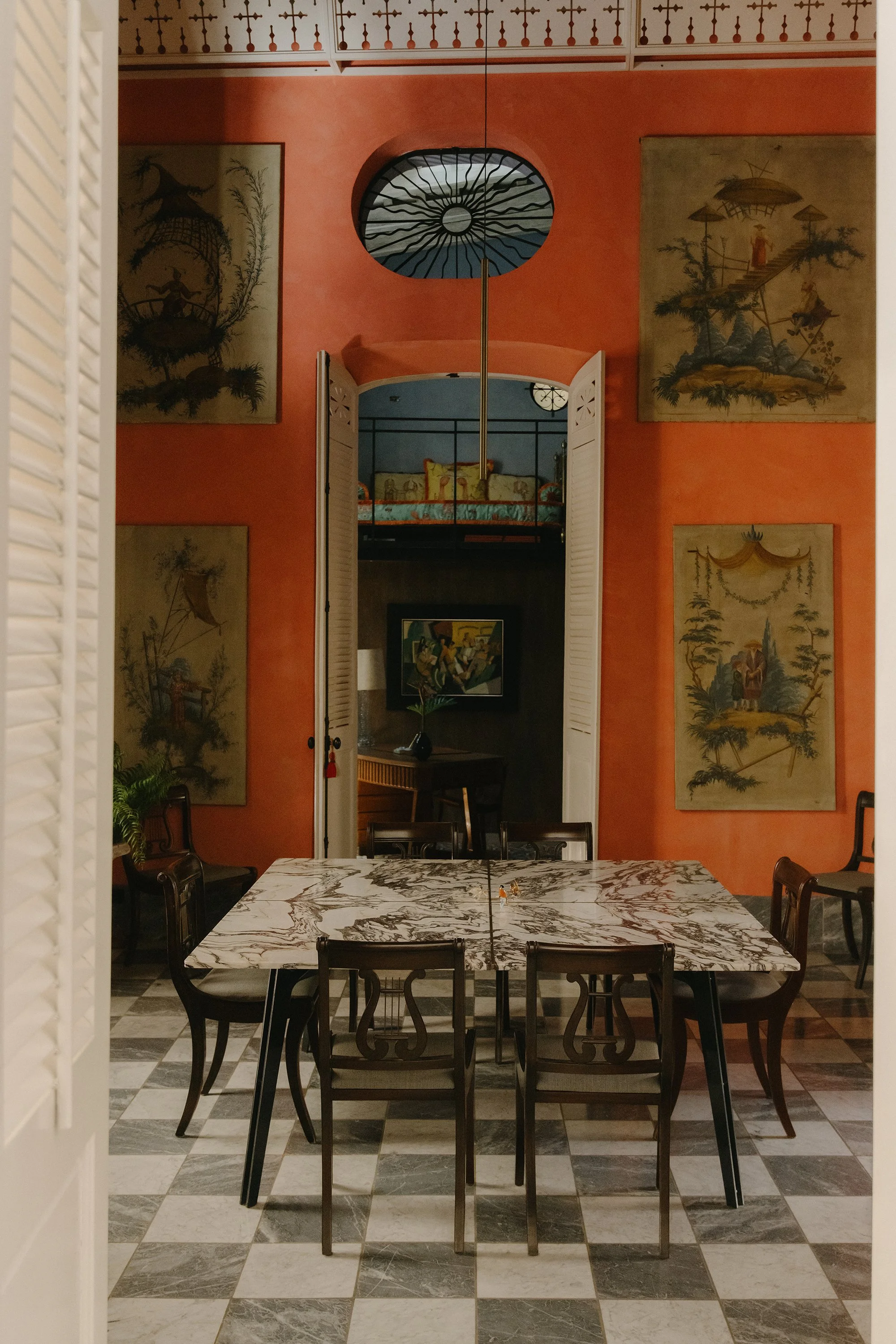 Dining room with a marble table, black chairs, orange walls, and artwork. A mirror reflects a room with a bed and a painting. White shutters are on the sides. Mundos Interiores. Ciudad Colonial de Santo Domingo.