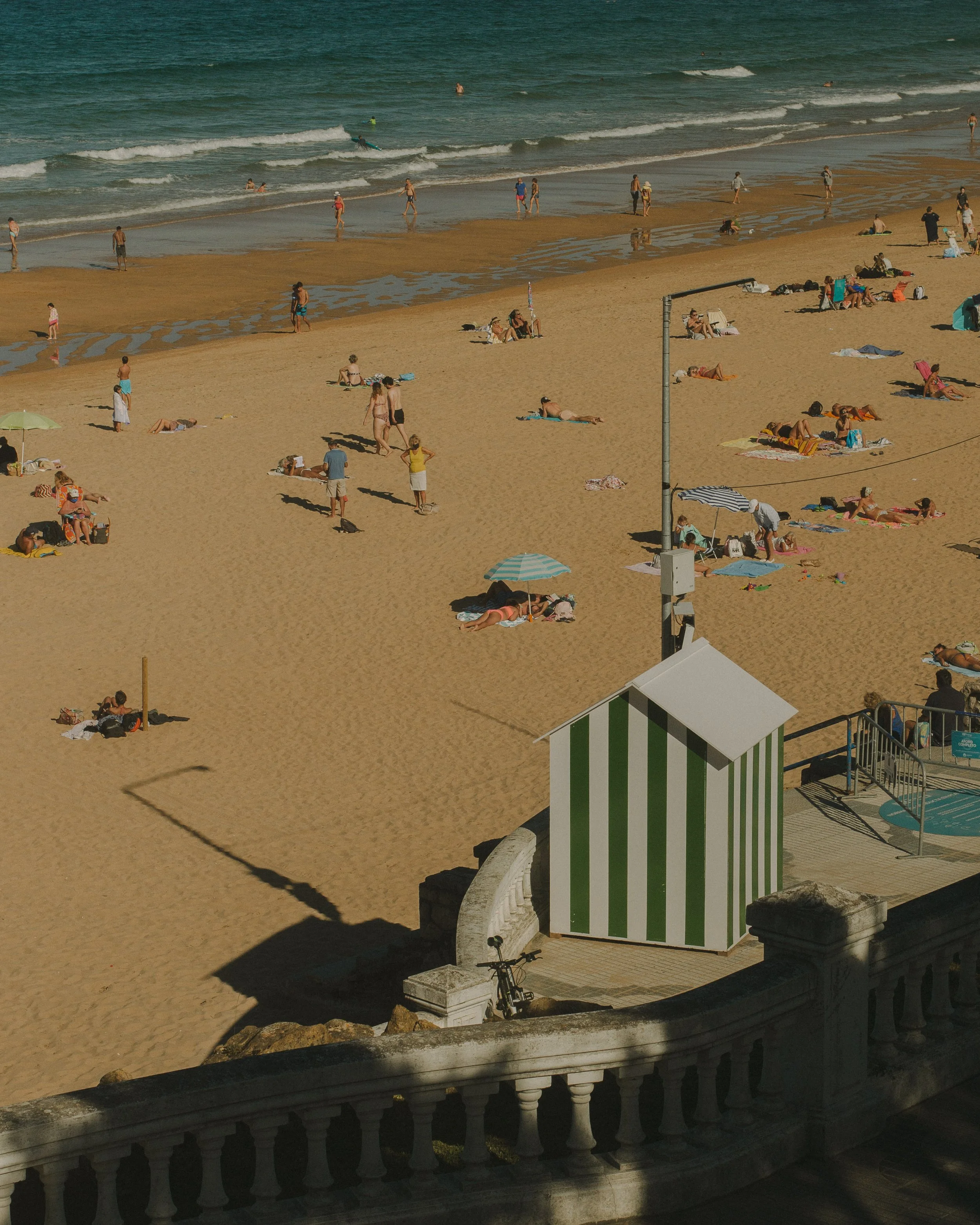 Beach scene viewed from a terrace, featuring people sunbathing and walking on the sandy shore, with ocean waves and swimmers in the background. A green and white striped beach hut and a bicycle are visible in the foreground.