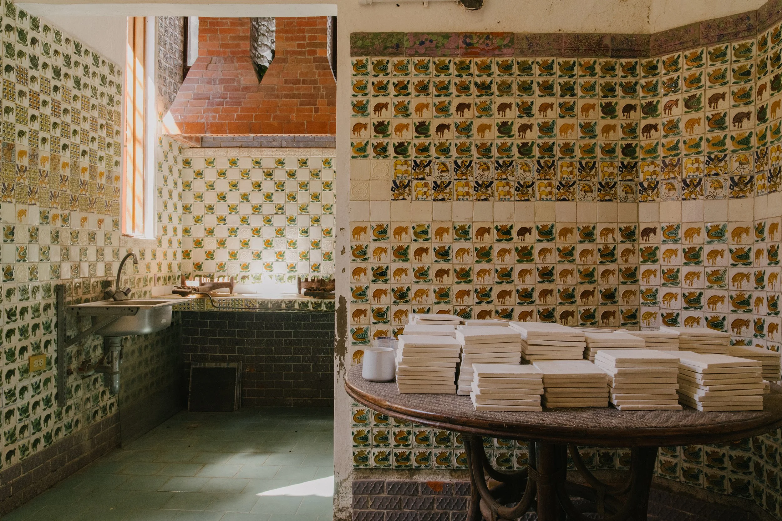 Interior of a room decorated with hand-painted tiles featuring animals and patterns, with a brick chimney, a window, and a table with stacked tiles. Mundos Interiores. Ciudad Colonial de Santo Domingo.