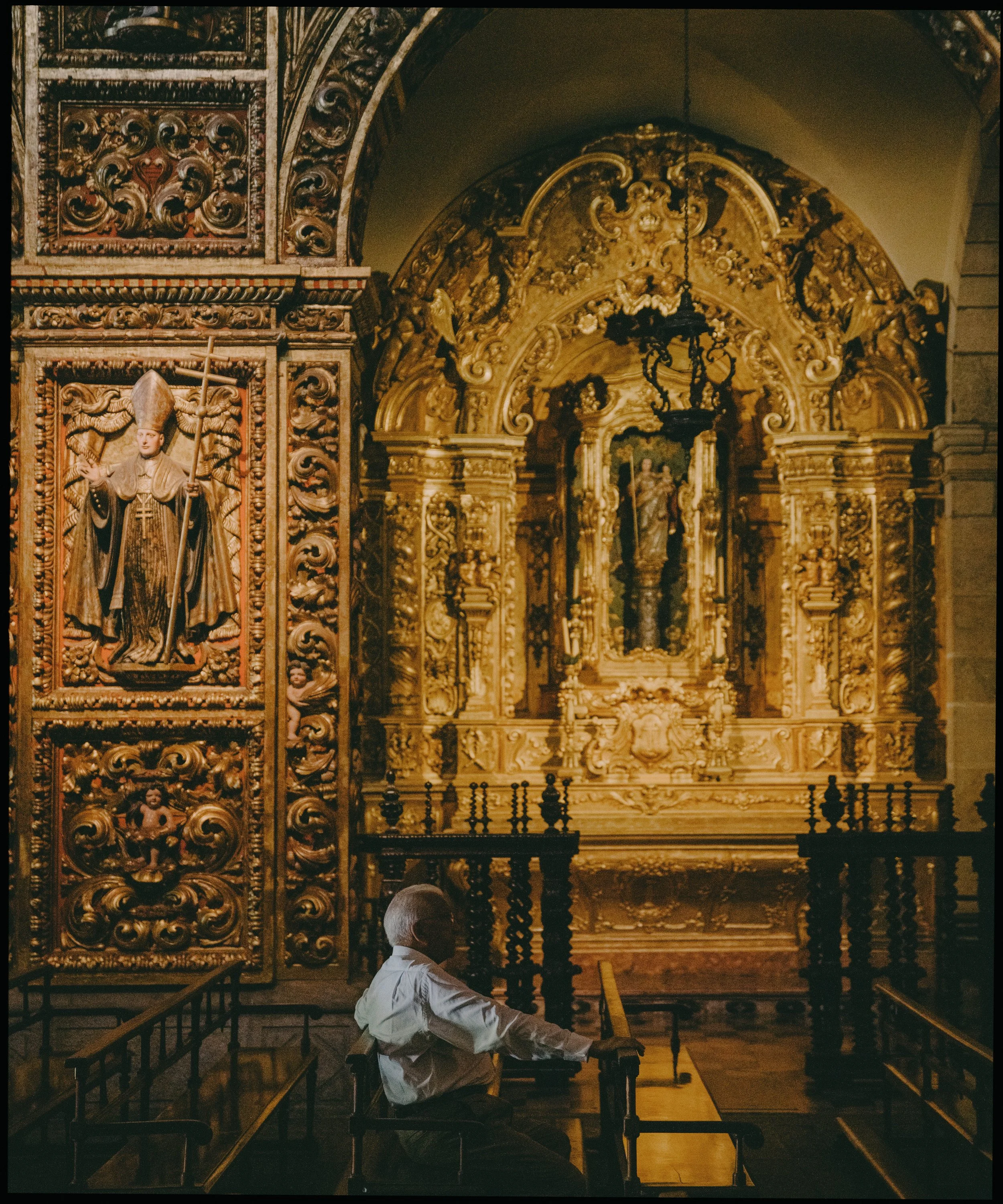 A man with dark hair, wearing a light-colored shirt, sitting in wooden pews inside an elaborately decorated church, with intricate gold carvings and religious artwork on the walls.