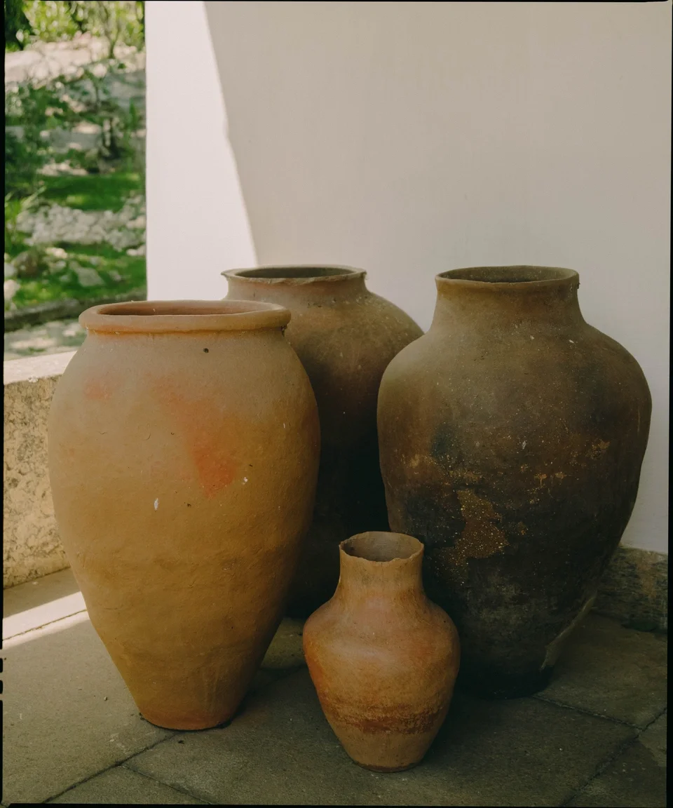 Four ancient clay pots of varying sizes and colors are arranged outdoors against a white wall, with a stone ledge and green trees in the background.