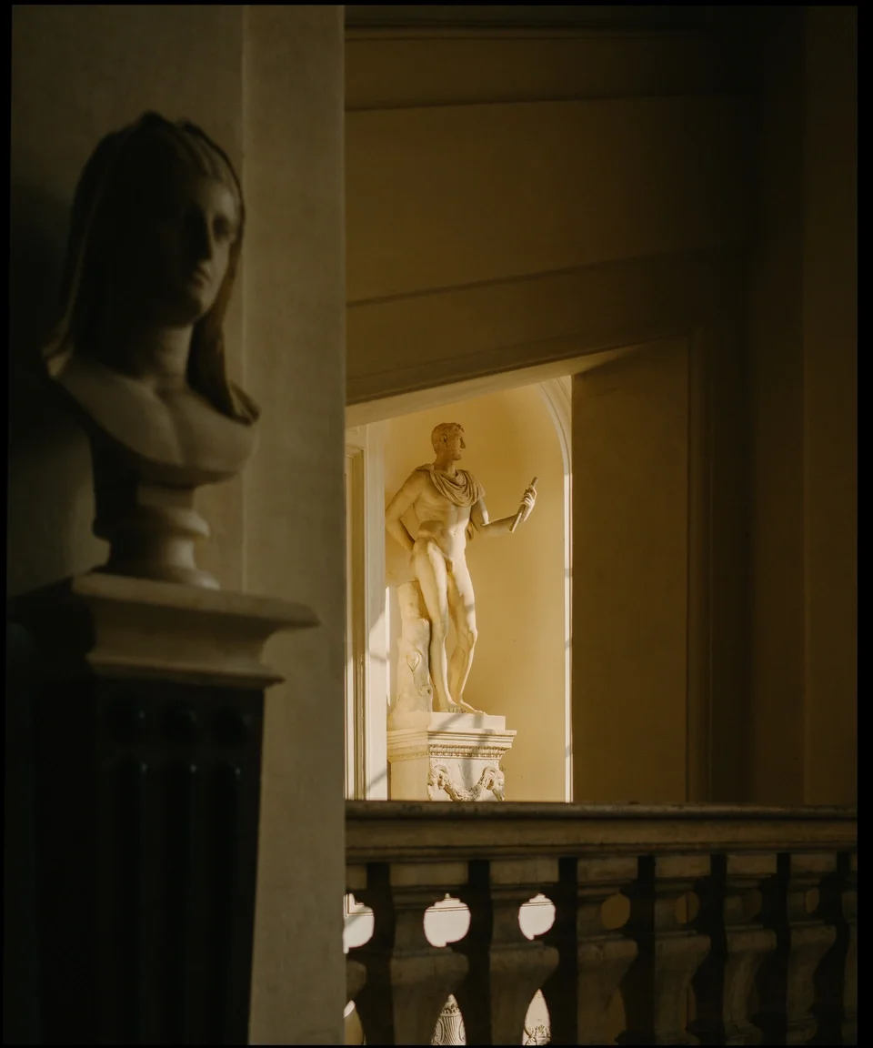 A classical sculpture of a man holding a scroll, viewed through an opening with a decorative balustrade, with another sculpture of a woman in the foreground.