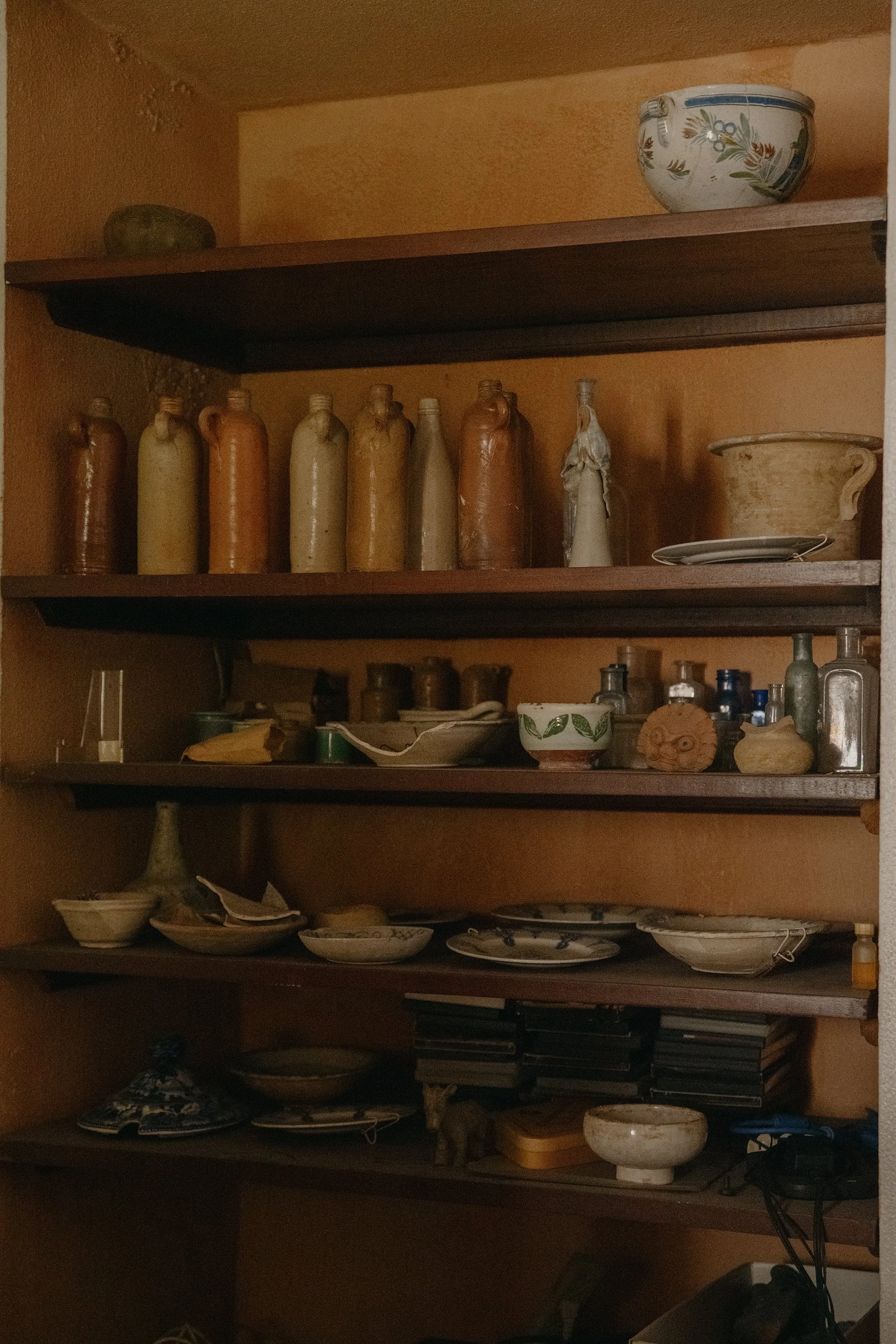 Wooden shelves filled with various ceramic and pottery items, including bowls, bottles, and vases, in a cozy indoor setting. Mundos Interiores. Ciudad Colonial de Santo Domingo.