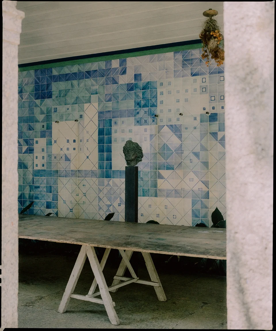 Photo of an interior space with a large mural of geometric blue and white tiles on the back wall, a wooden table in the foreground, and a statue of a head on a pedestal in front of the mural.
