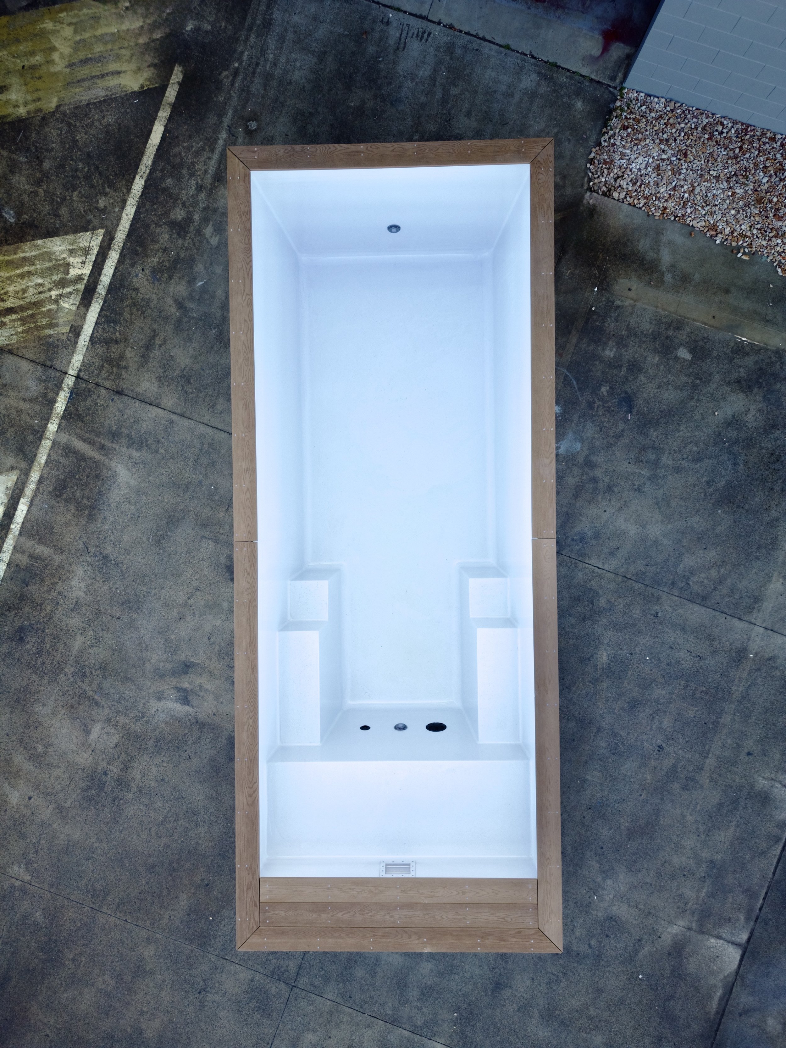 Empty shower stall with a white interior, a drain, and plumbing fixtures at the bottom, framed with a wooden border, on a concrete floor.