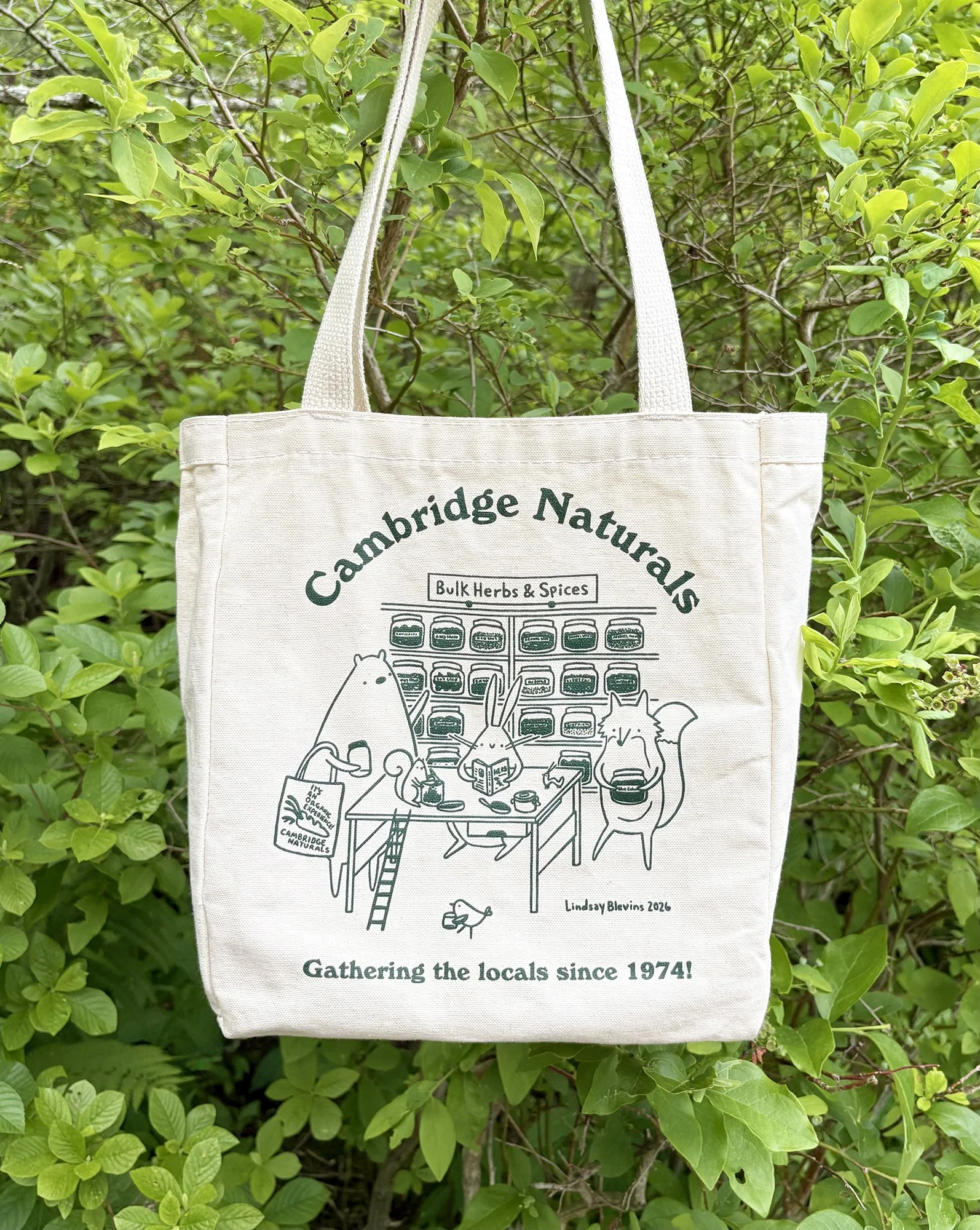 A canvas tote bag for Cambridge Naturals with an illustration of woodland animals gathered in the store's bulk herb section. "Gathering the locals since 1974" is printed below the illustration.