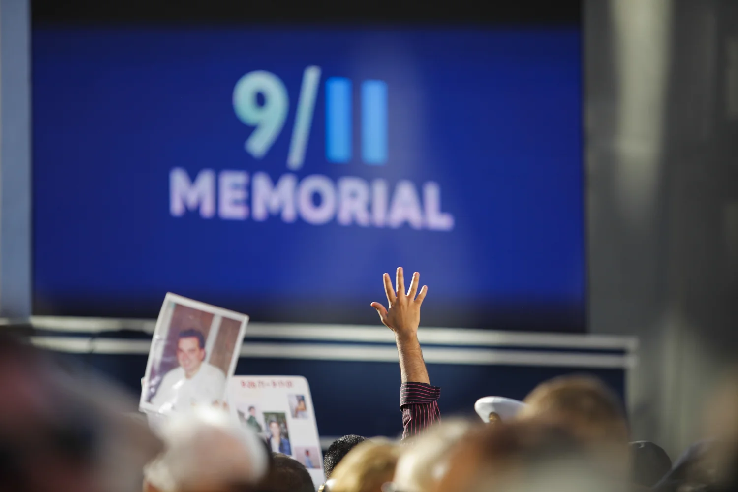 People hold up signs and photos, with an individual raising a hand towards the sky in the crowds at the Sept. 11 10th Anniversary Commemoration Ceremony at Ground Zero in New York, Sept. 11, 2011.