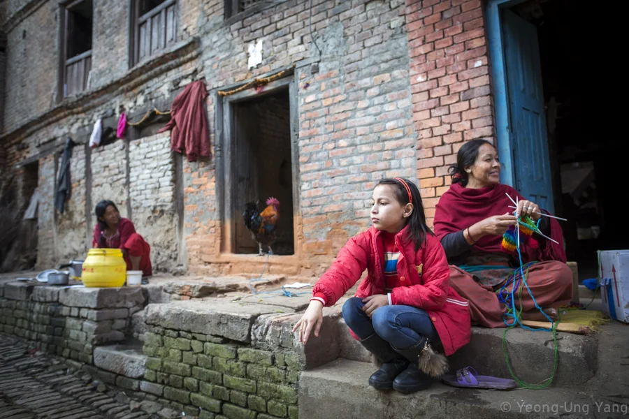 A youth throws a rock on the streets of Bhaktapur, Nepal while her mother is hand-knitting, November 22nd, 2014. According to the Ministry of Labour and Employment, more than 520,000 labour permits were issued for foreign employment in the 2013-2014 fiscal year. Many youths also prefer to work abroad.