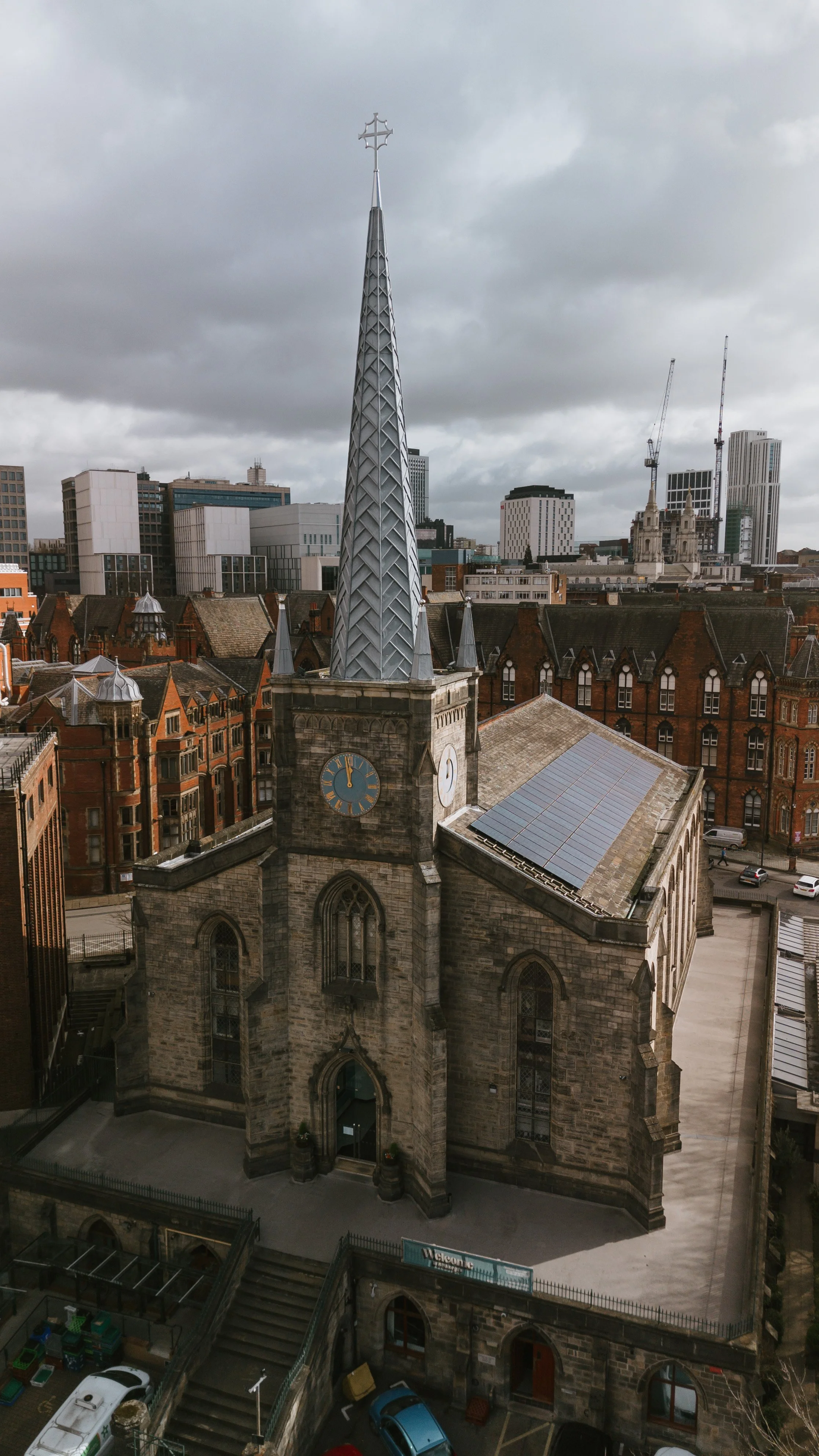 A tall stone church with a clock tower, topped with a modern metallic spire and cross, in an urban setting with high-rise buildings and construction cranes in the background.
