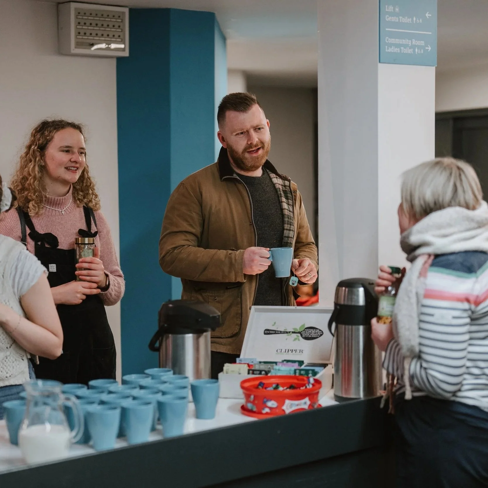 People gathered around a coffee station at an indoor event, with coffee cups, a tea box, and a thermal coffee pot on the table, engaging in conversation.