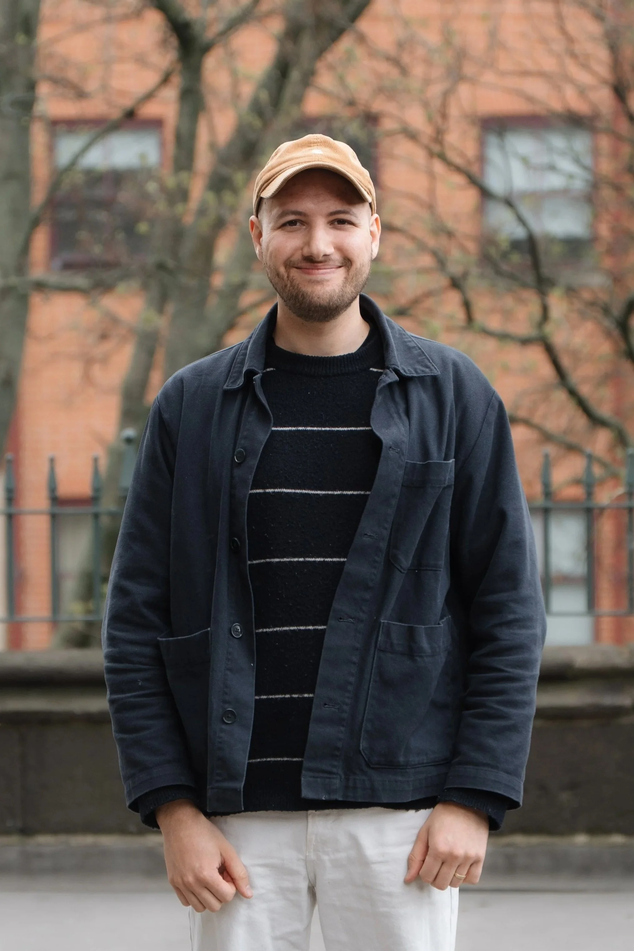 A young man with a light beard and smile, wearing a tan cap, black sweater with white stripes, dark jacket, and light pants, standing outdoors in front of a background with a brick building and leafless trees.