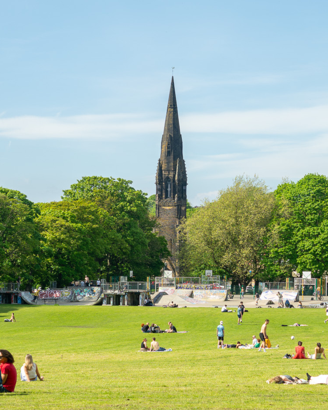 View of a church steeple rising above trees, with a park in the foreground where people are relaxing and playing.