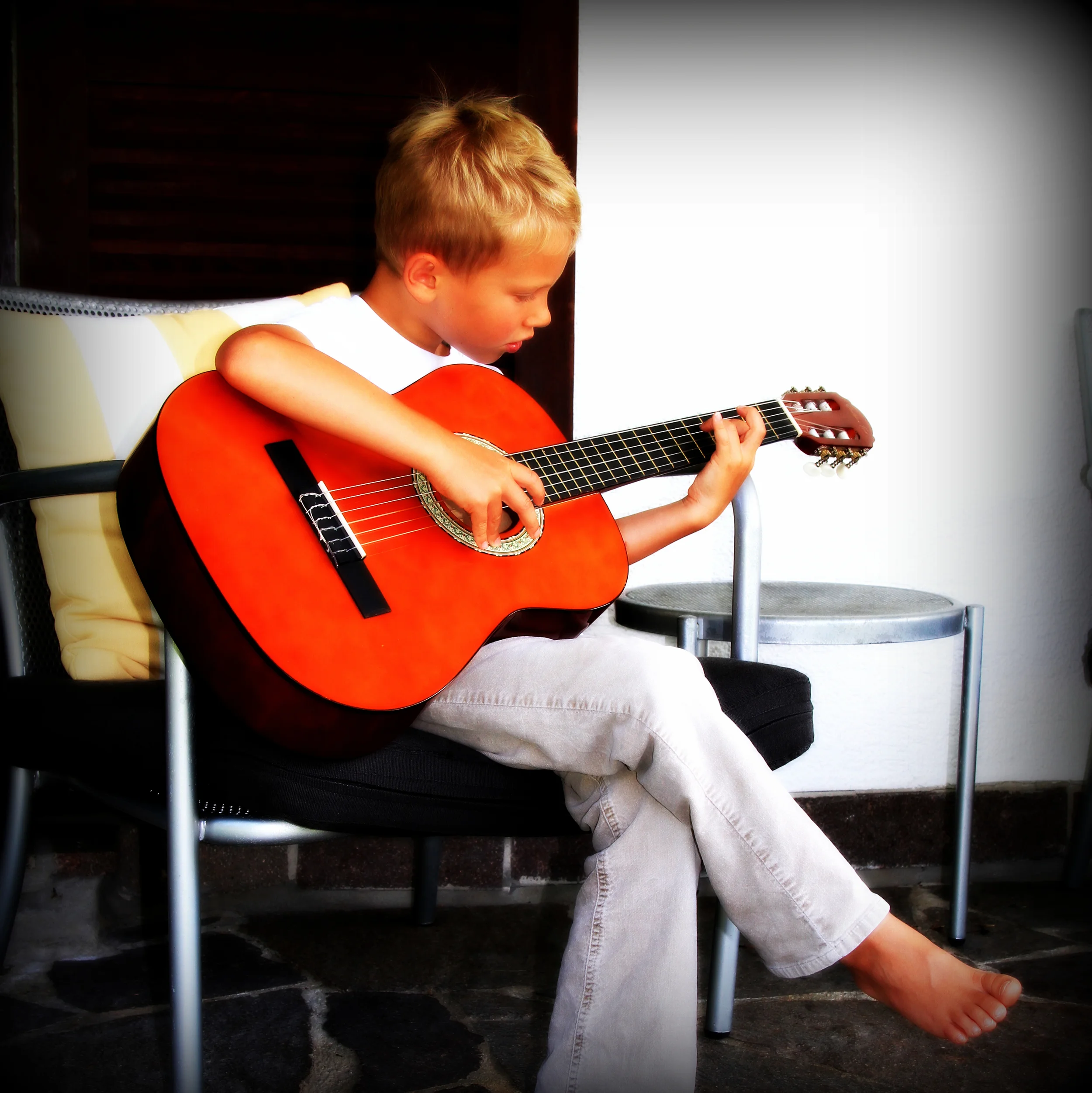 A young boy with blond hair sitting on a chair, playing a red guitar | Boulder Guitar Coach | Guitar Coach for beginners
