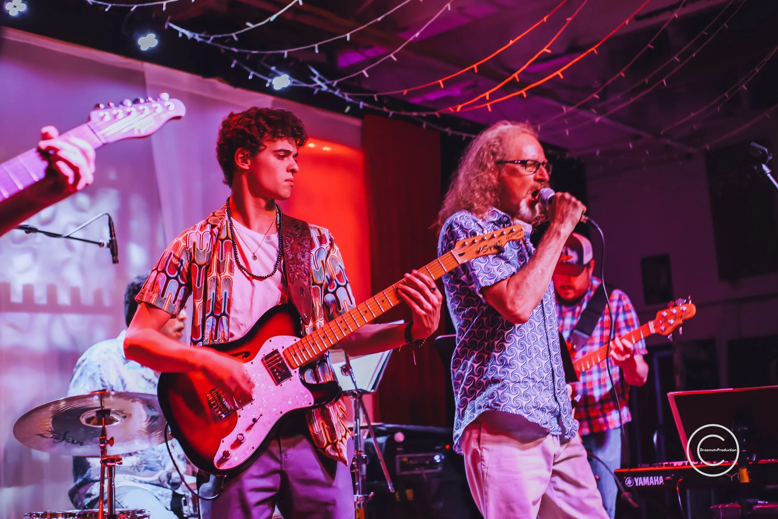 A band performing on stage with purple and red lighting. The band includes a young man playing an electric guitar, an older man singing into a microphone, and other musicians playing instruments such as drums and bass. The stage is decorated with colorful string lights.