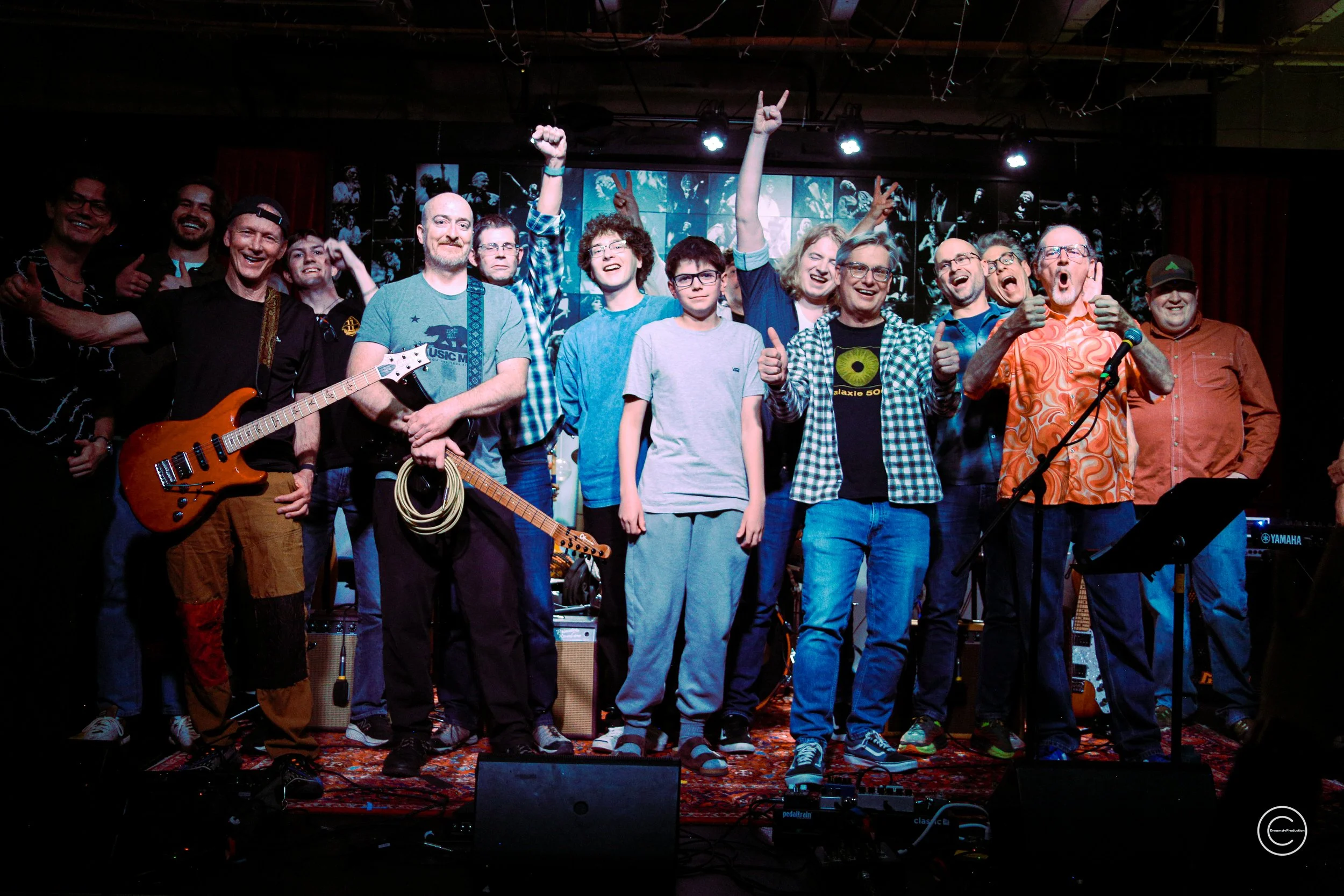 Group of people on stage celebrating, some holding musical instruments, with a multicolored light display in the background.