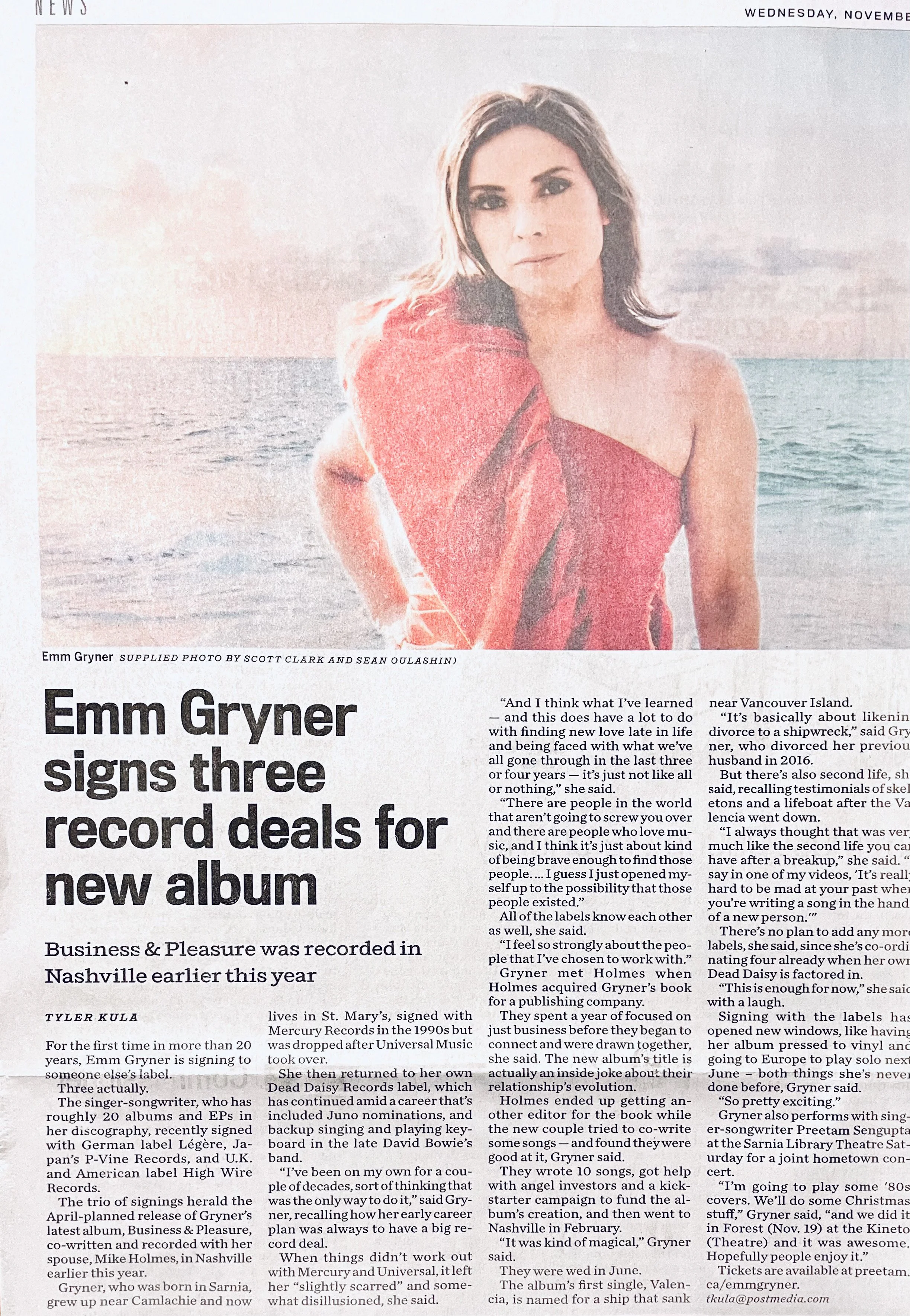 Emm Gryner is wearing a red strapless dress, standing near the ocean during sunset.