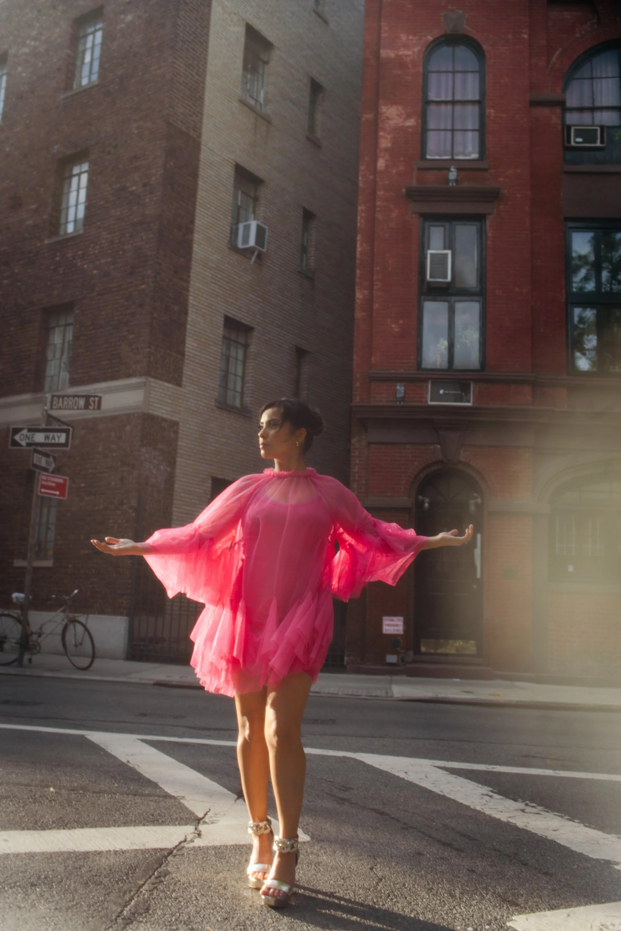 Emm Gryner is wearting a flowy dress and high heels, standing on a crosswalk in front of brick buildings in an urban setting, with sunlight shining on her.