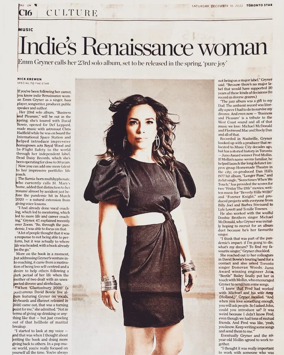 Emm Gryner is wearing a black dress with a cutout at the waist, standing in front of a newspaper article about her.