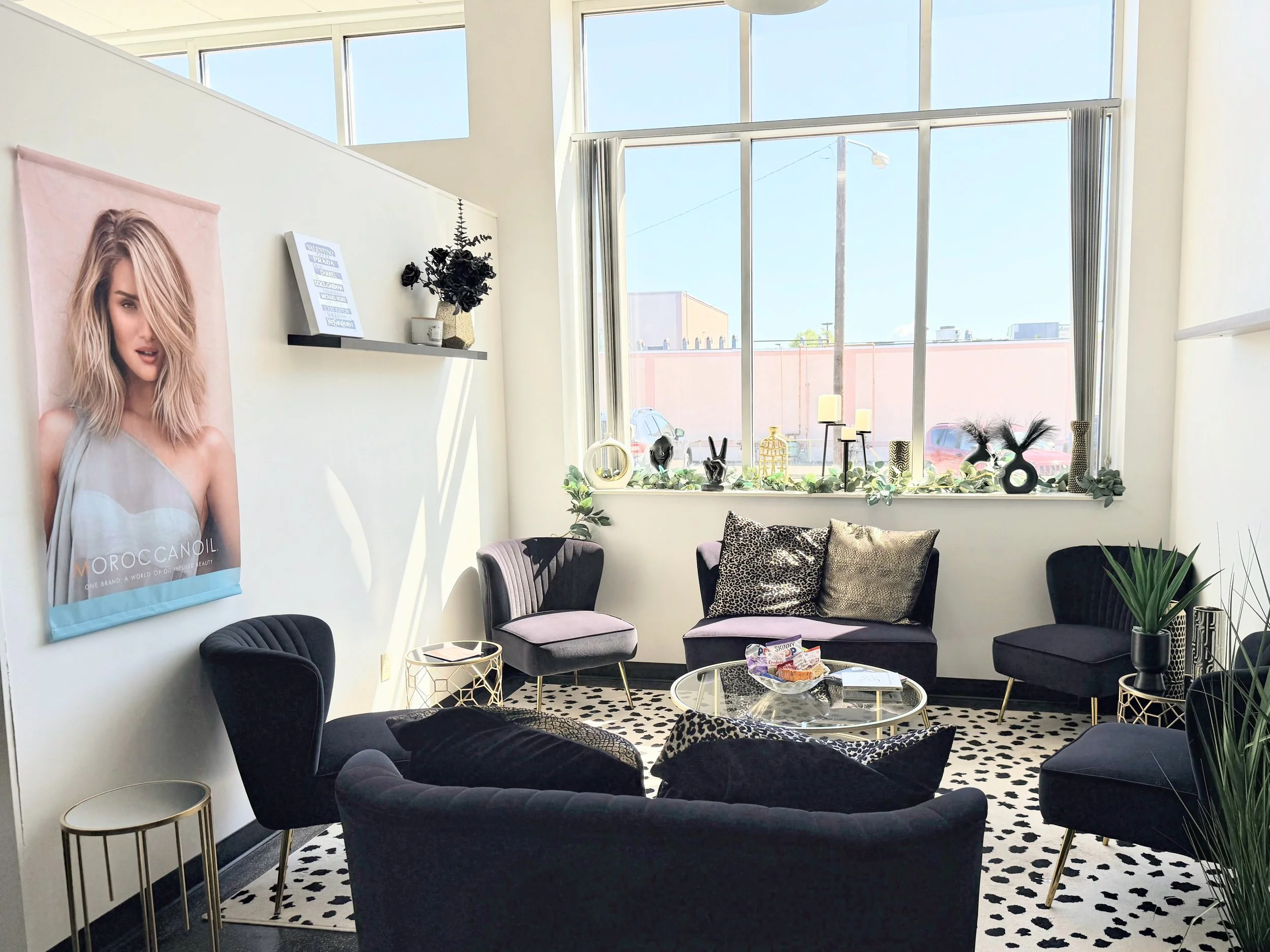 A modern waiting area with four black velvet armchairs, gold side tables, and decorative black and leopard print pillows. Large windows let in natural light, and the sill is decorated with plants, candles, and black decorative objects. A leopard print rug covers the floor, and a poster of a woman with blonde hair and a grey top is on the wall.