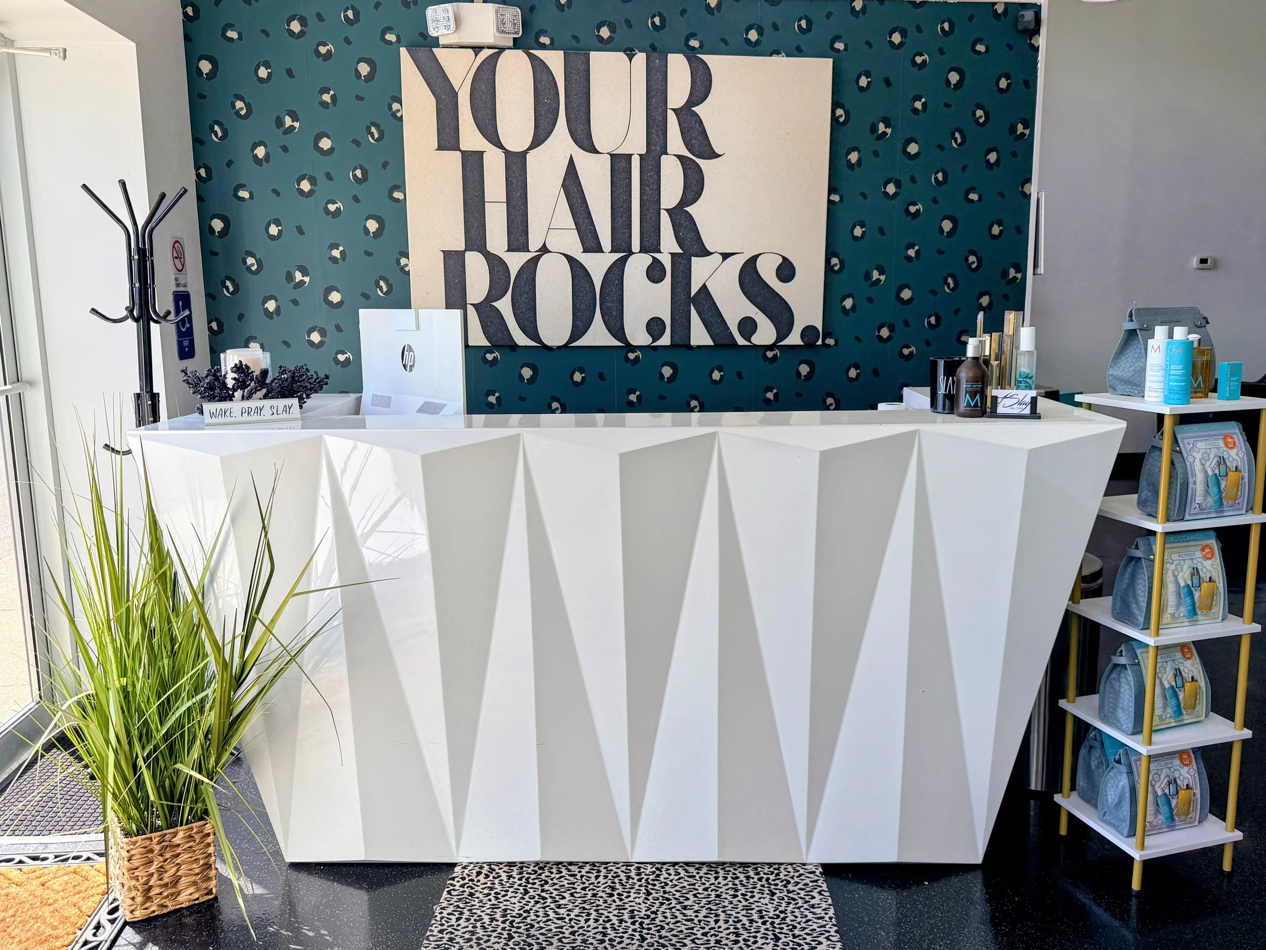 Interior of a hair salon or beauty shop with a large sign that reads "Your Hair Rocks" on a patterned wall. There is a white, geometric reception desk in front of the sign, a small table with hair products and accessories, and a shelving unit with hair care items on the right. A basket with purple flowers and a green plant are visible in the foreground.