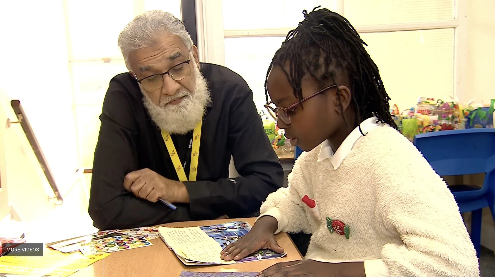 Man sits with girl at a school table listening to her reading a book