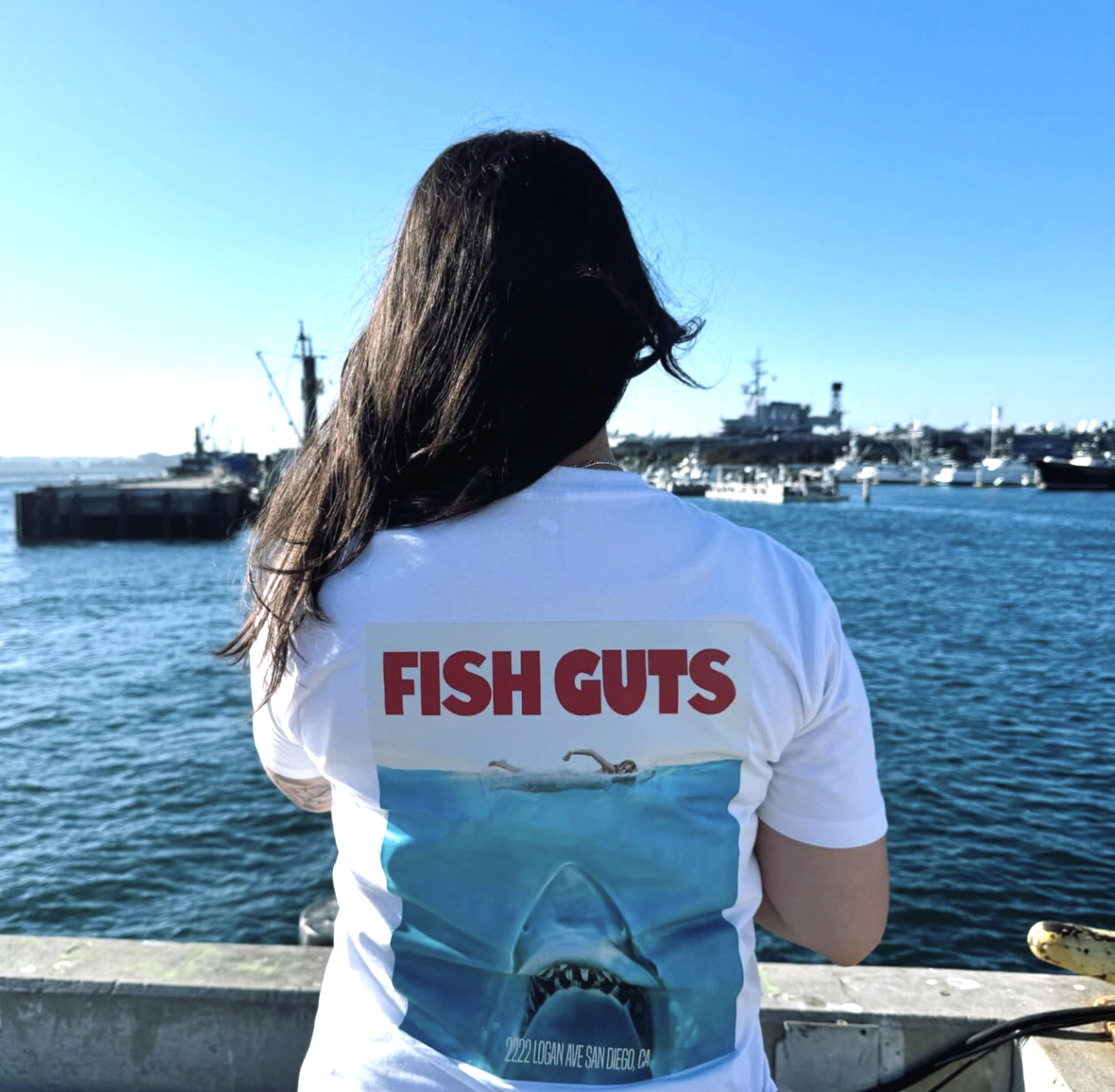 Person with long dark hair wearing a white t-shirt with red text on a boat dock near water with ships and marina in background.