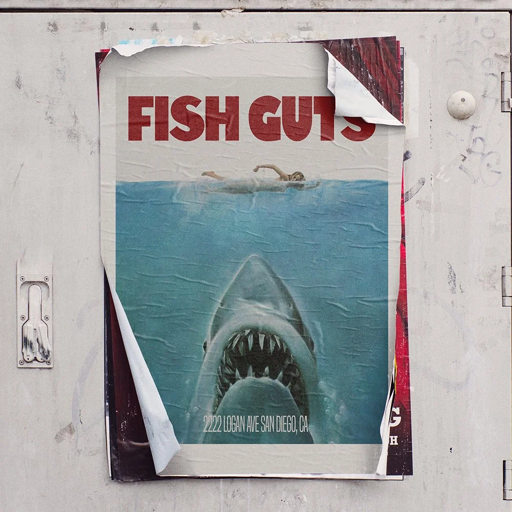Poster titled "FISH GUTS" depicting a swimmer and a large shark underwater with its mouth open, showing sharp teeth, and an address at the bottom. The poster is partially torn at the top right corner.