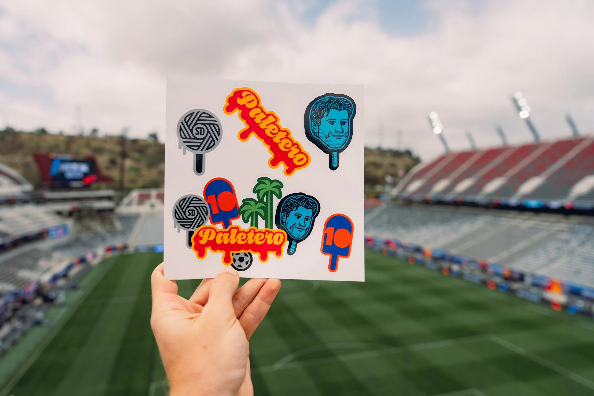 Hand holding a sticker sheet with various stickers including the word 'Championship', a face on a popsicle, palm trees, and animals, with a sports stadium in the background.