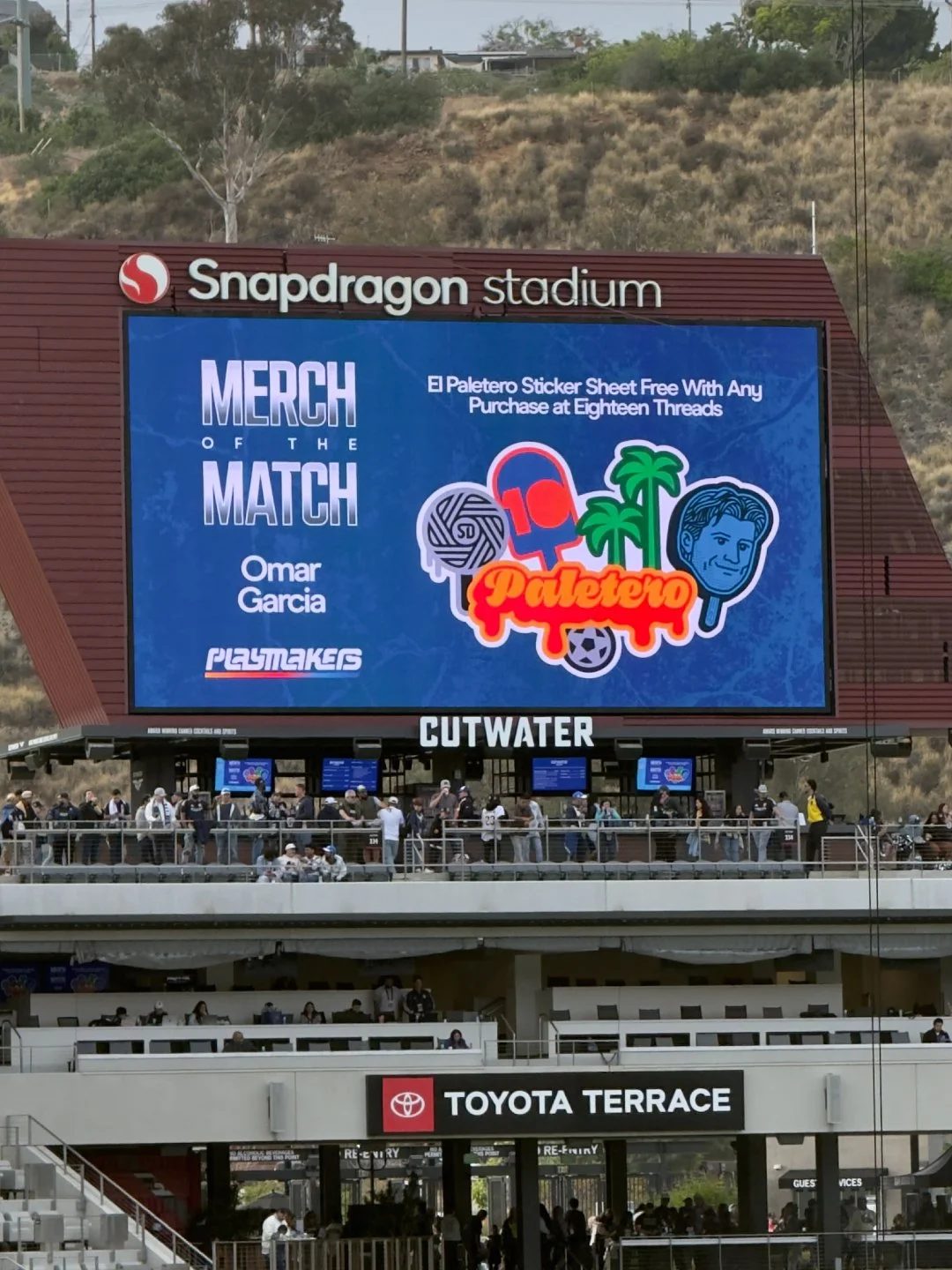 Large digital screen at Snapdragon Stadium promoting the movie 'Merch of the Match' featuring Omar Garcia and Playmakers, with a special offer for free Palatero sticker sheets at Eighteen Threads. Below, spectators gather at the stadium's seating area and concourse, with signage for Toyota Terrace and Cutwater visible.