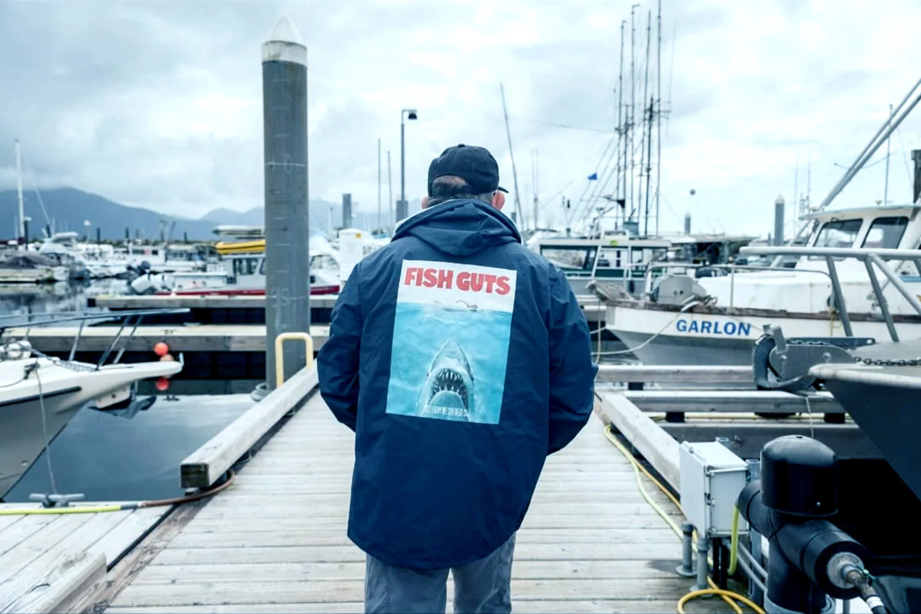 A person walking on a dock at a marina with boats, mountains in the background, and cloudy sky. The person is wearing a jacket with 'FISH GUTS' printed on the back and a shark image.