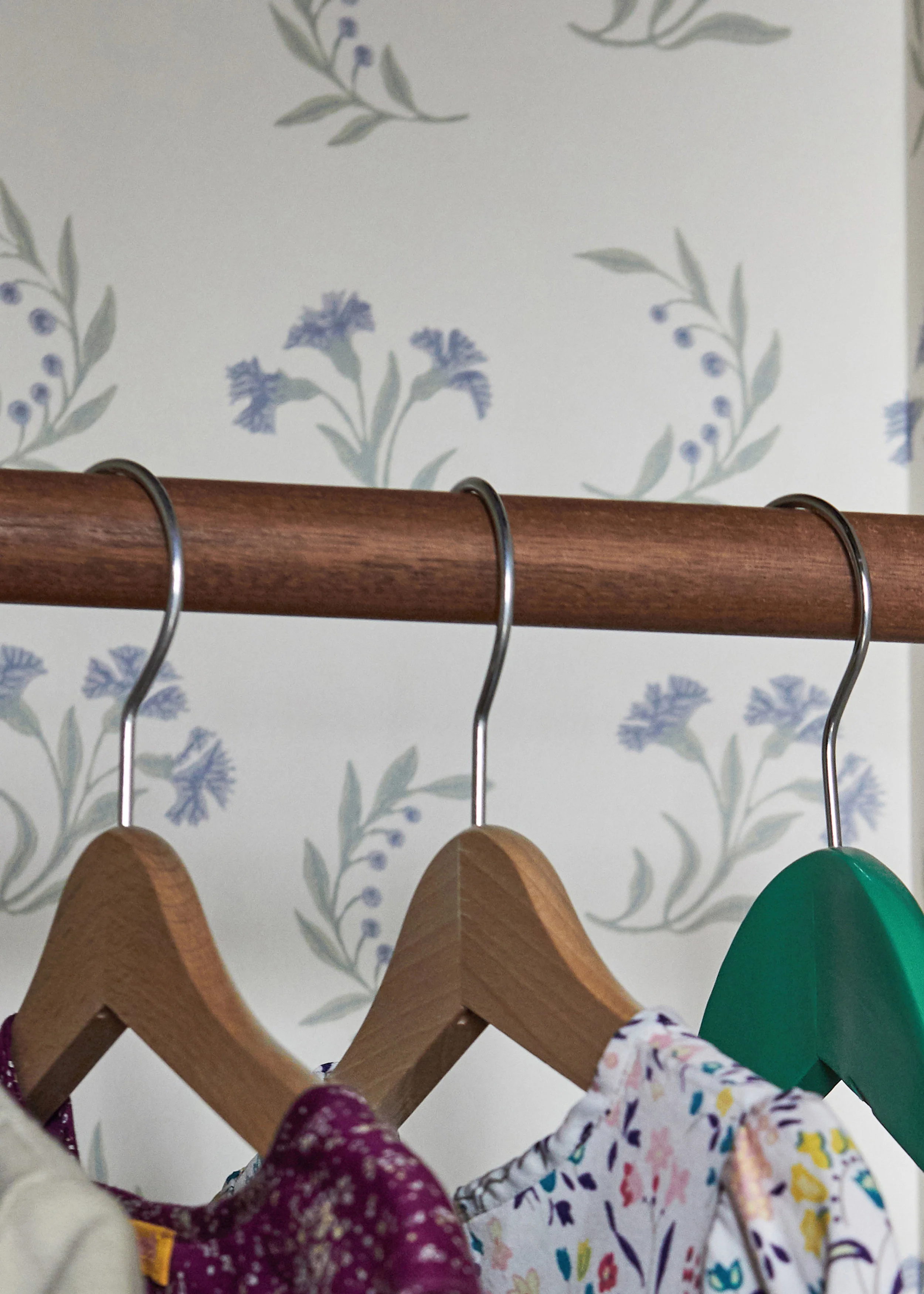 Sapele Hardwood Wardrobe Rail - Close-up of a brown waxed timber rail with three hangers and dresses, set against floral wallpaper by Robert Kime. Made from sapele hardwood.