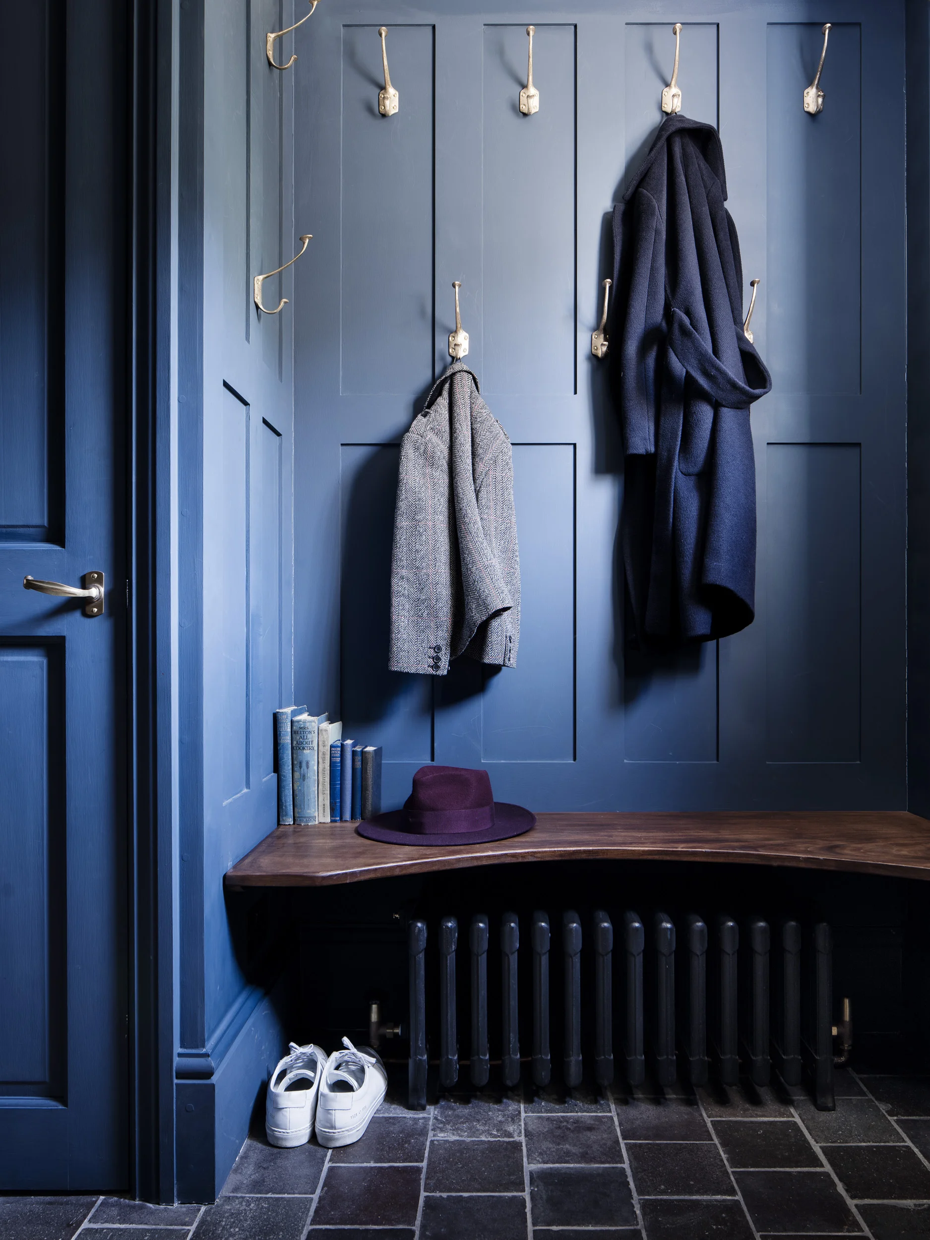 Nine Jamie Coat Hooks in cast bronze mounted in a navy blue paneled hallway alcove, with a bench seat below.