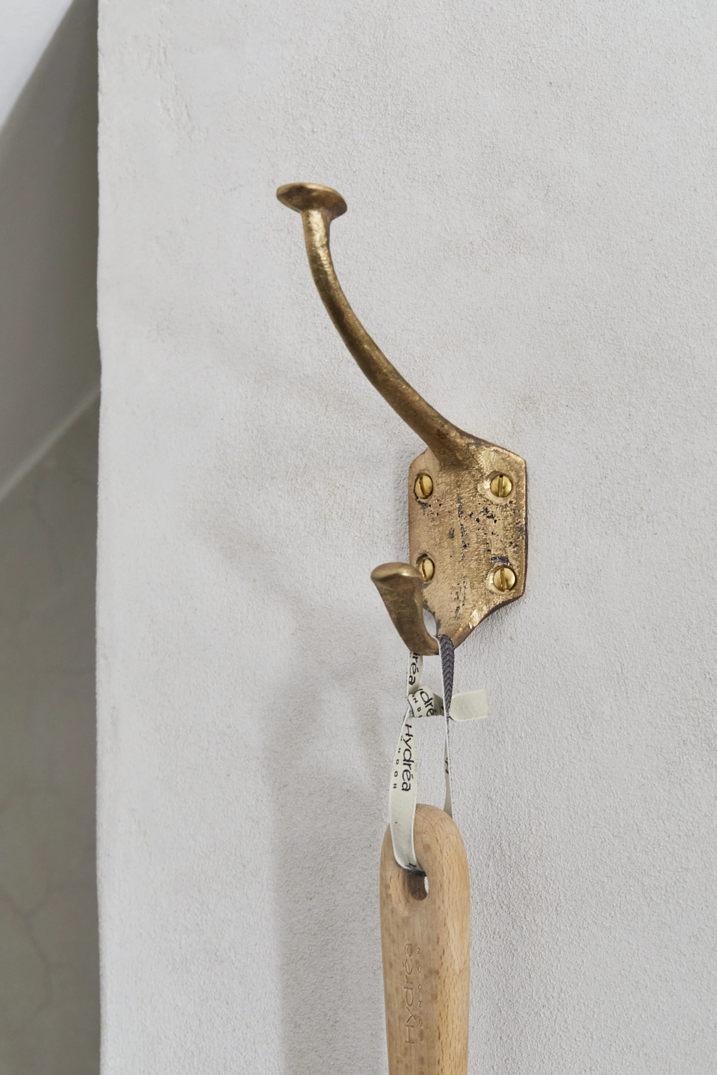 Close-up of the Jamie Coat Hook in cast bronze, fixed to a lime plaster wall. The texture of the sand casting on the hook and the lime plaster surface are clearly visible.
