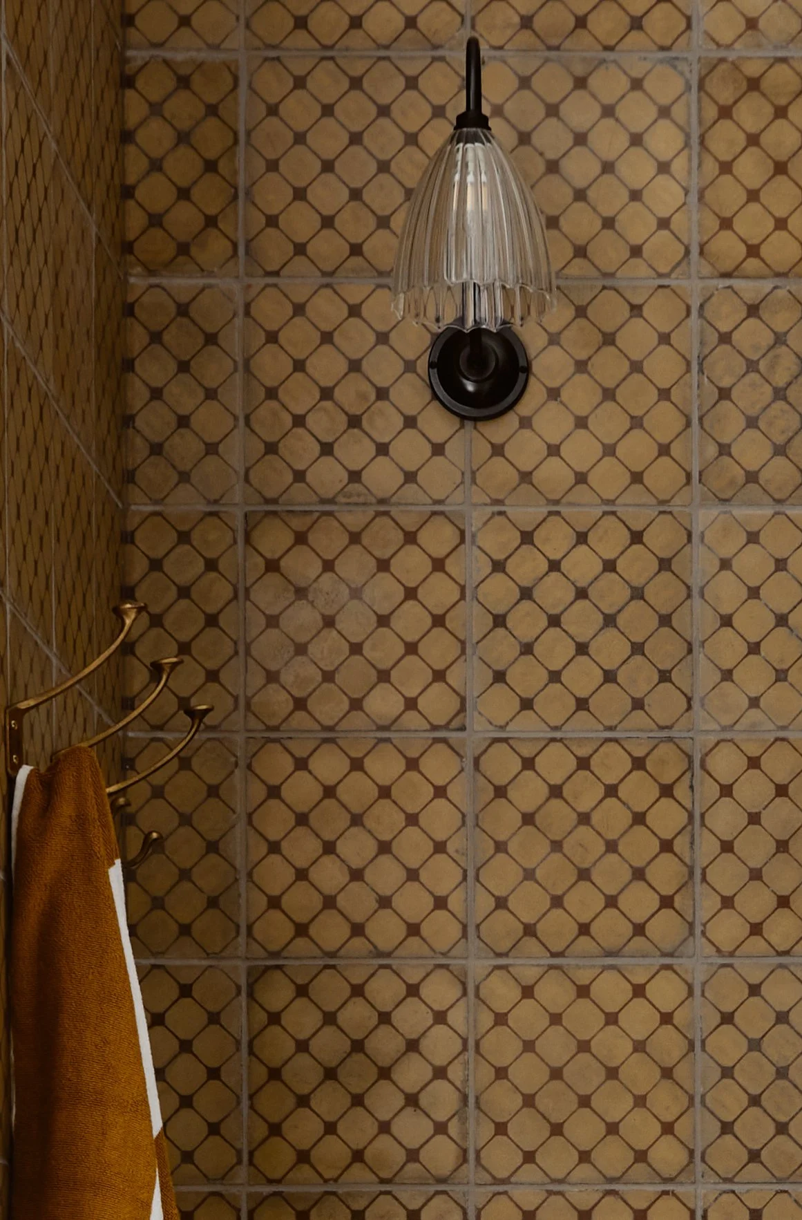 Side view of three Jamie Coat Hooks in cast bronze in a shower area with Spanish encaustic tiles. A bronze wall light is visible in the background.