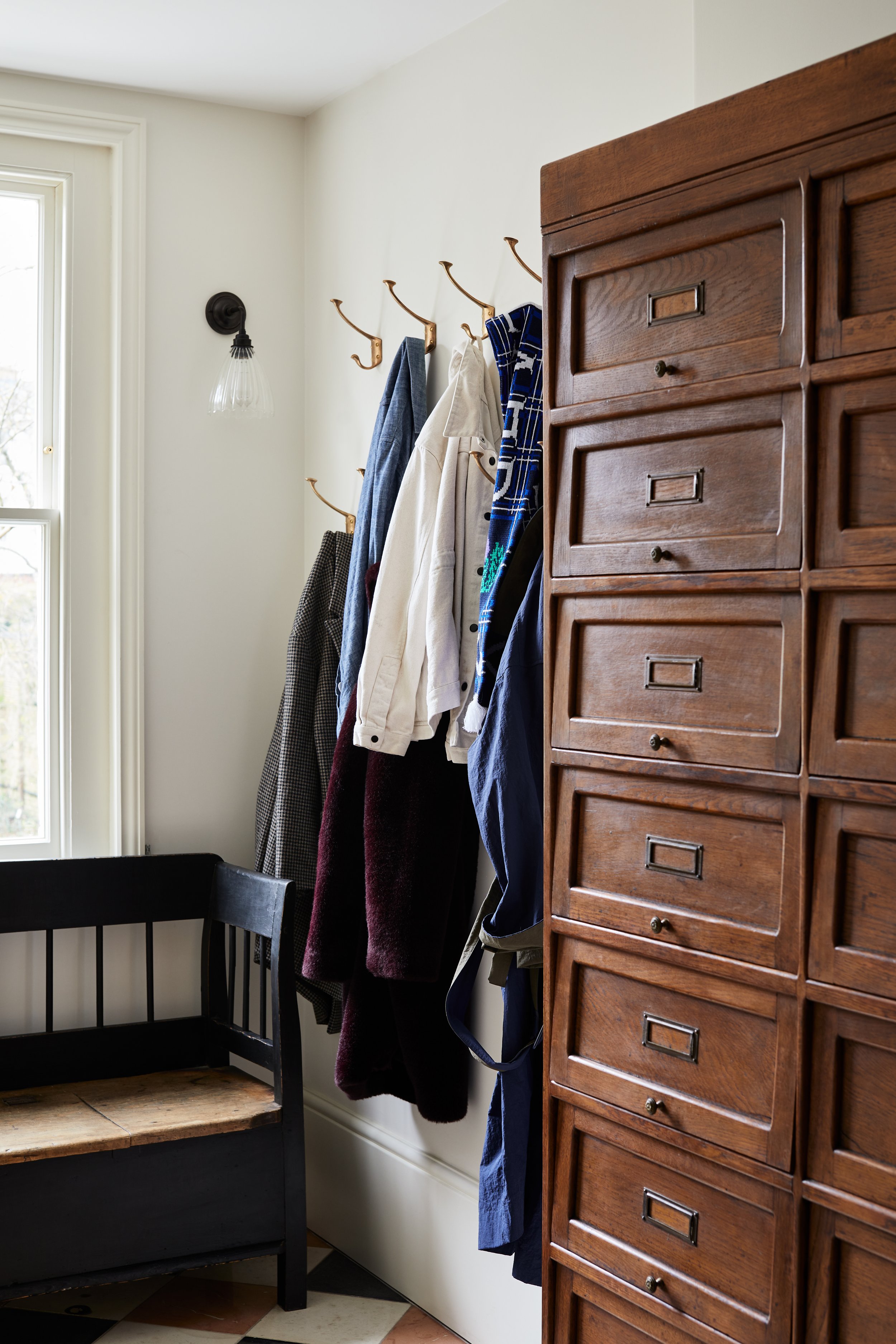 Jamie Coat Hook in cast bronze mounted in a white hallway with coats hanging. A bench seat is to the left, with a pigeonhole storage unit in the foreground.
