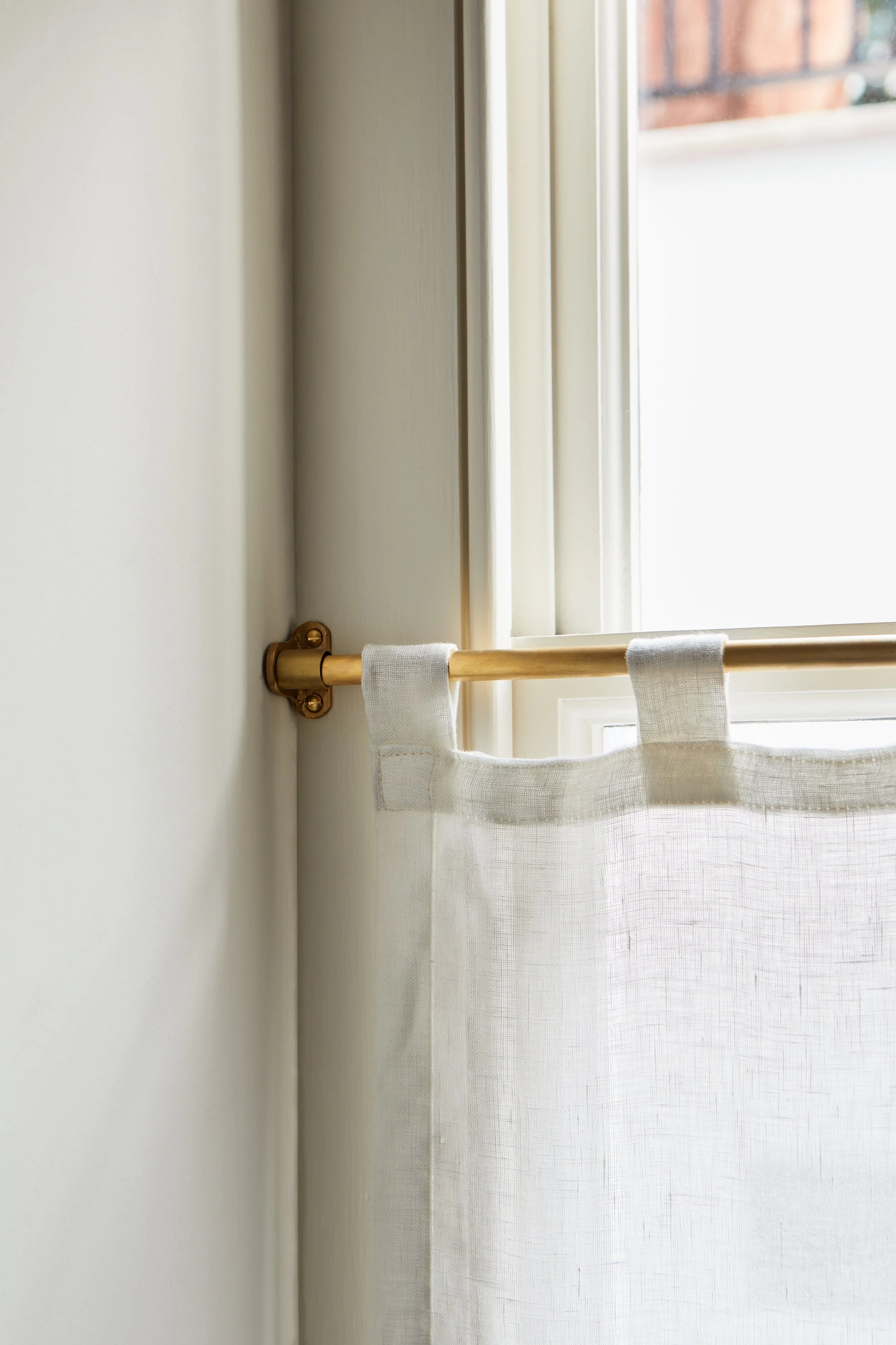 Café Curtain End Brackets - Wider shot of the bracket fixed to a window frame, with the cross-hatch texture of fabric visible. Made in raw brass.