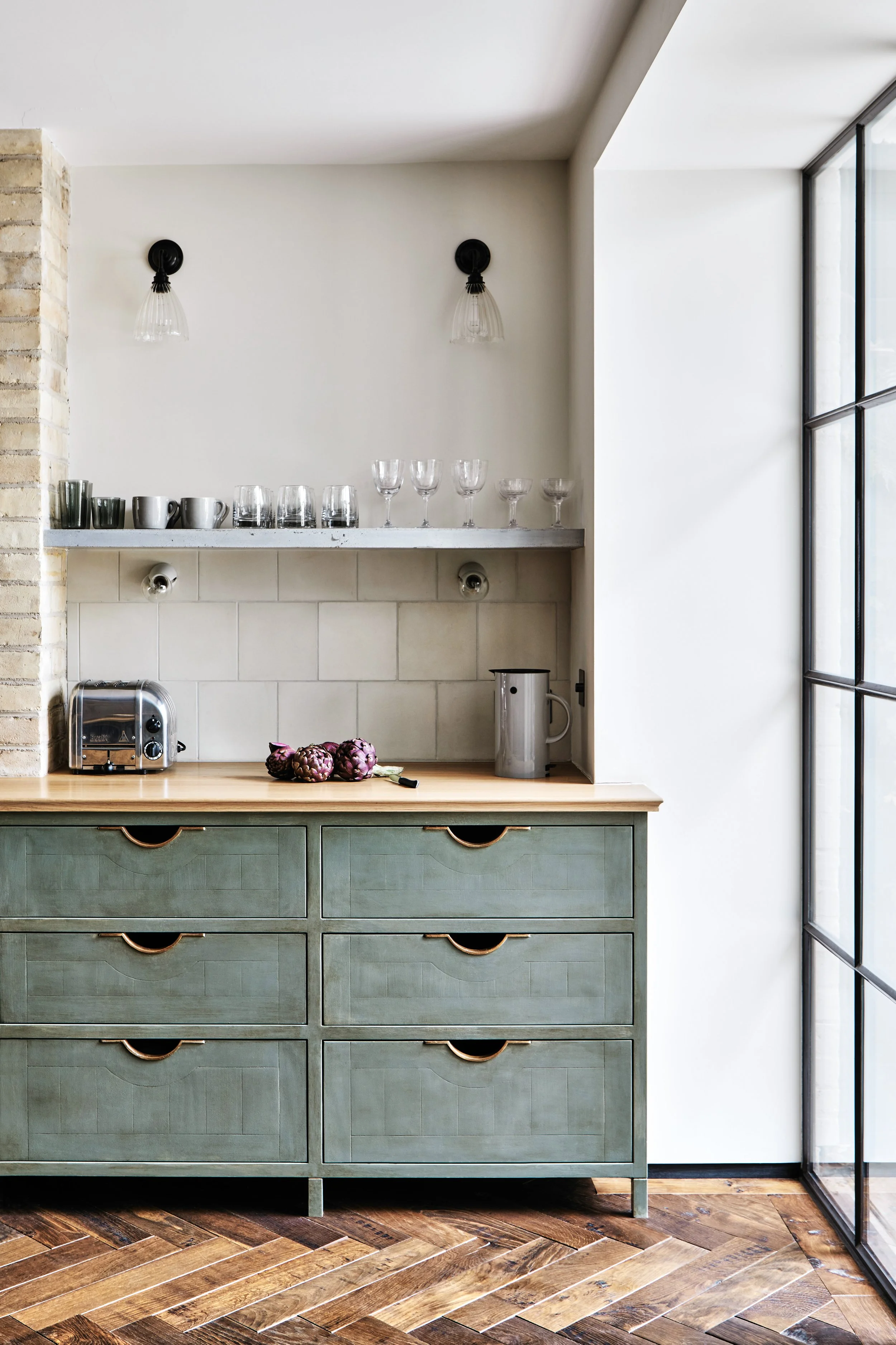 Mid-shot of a bank of 6 drawers with Moray drawer pulls in a painted green kitchen, featuring an oak surface, parquet floor, and a concrete shelf above with glasses on it.